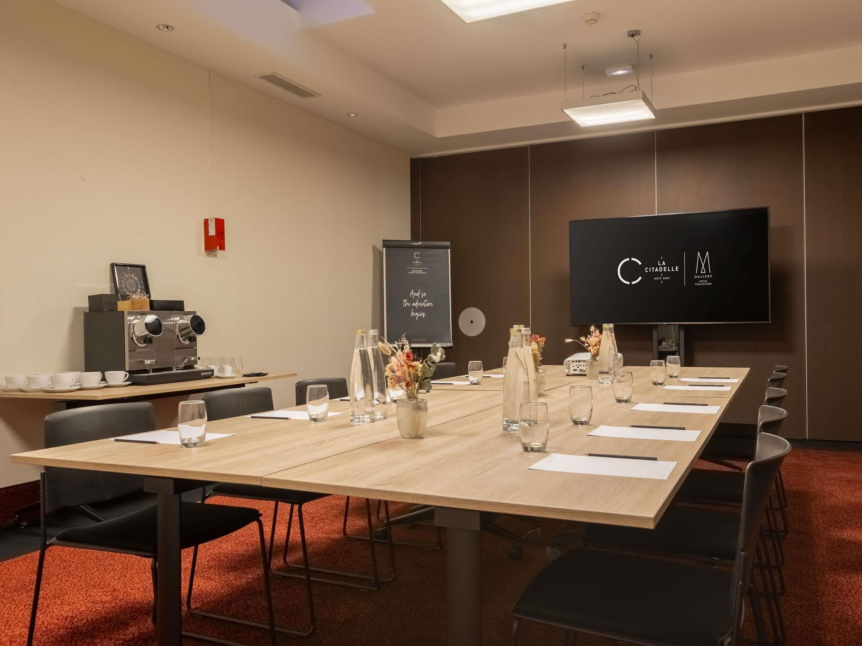A conference room with a long table, chairs, a projector screen, a coffee machine, and a TV displaying a logo.