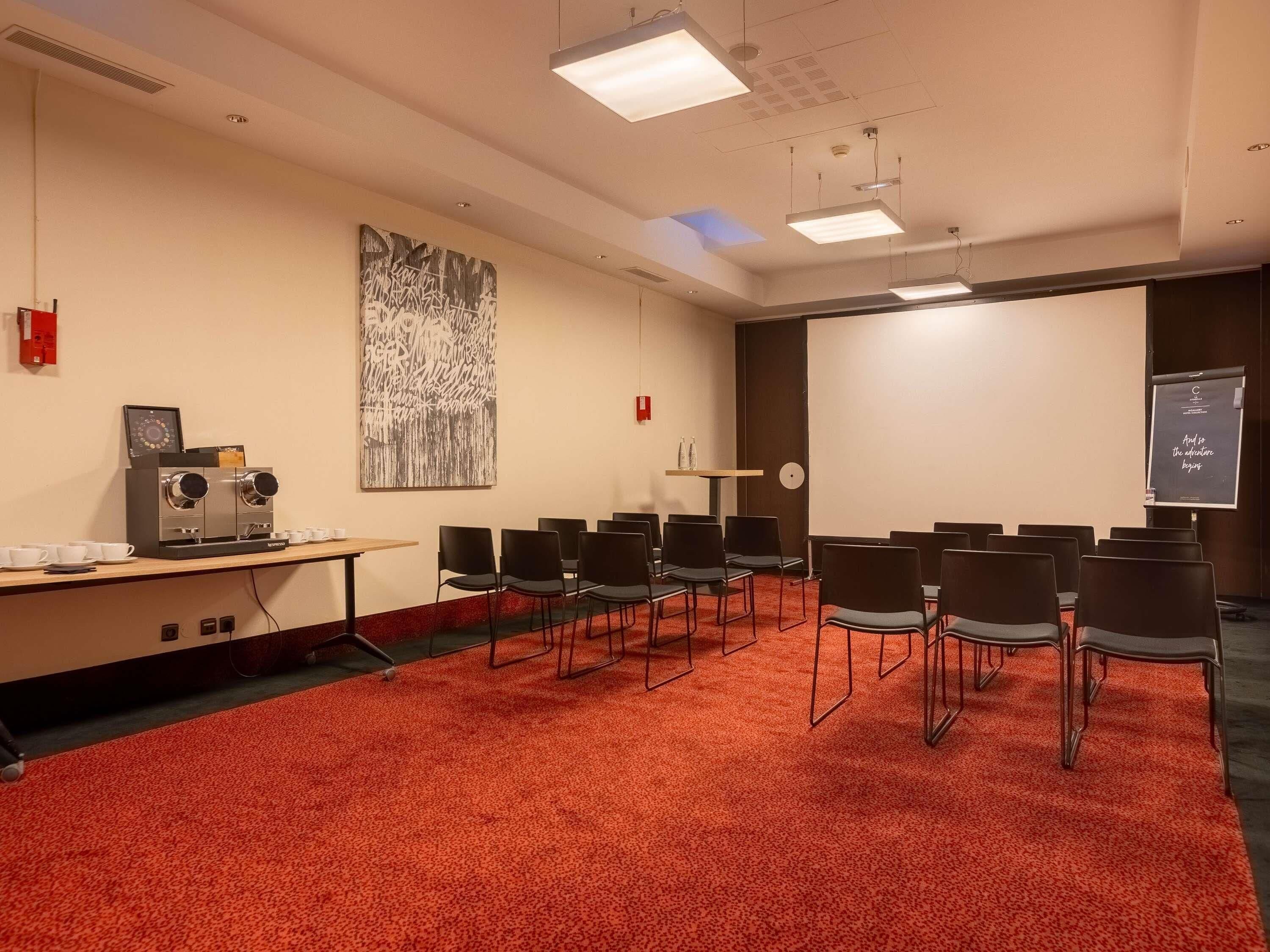A conference room with a red carpet, a coffee machine, a projector screen, and a large abstract painting on the wall.
