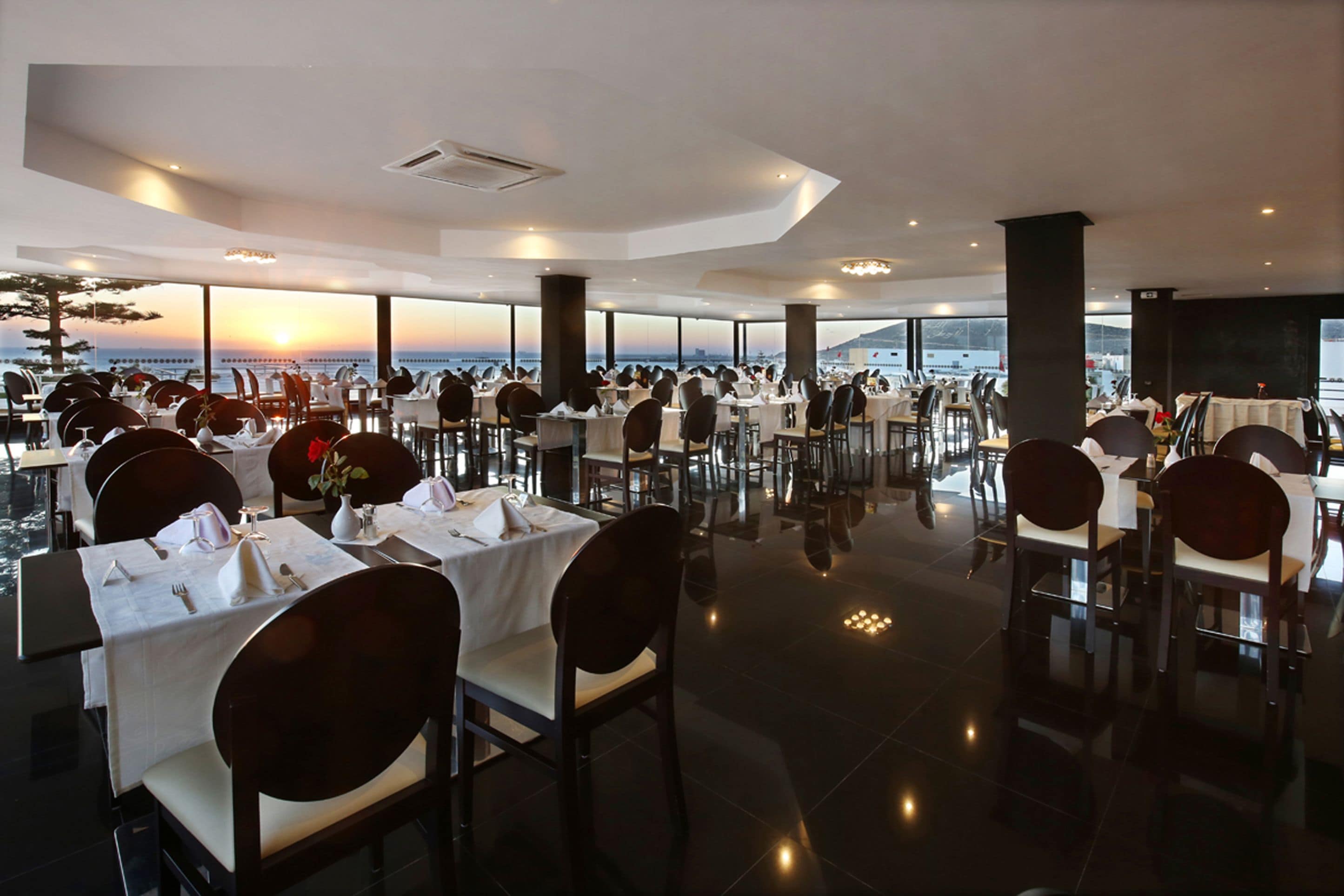 Un espaiós restaurant amb taules preparades per dinar, grans finestres que ofereixen vista al mar, i un disseny interior modern.