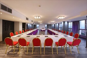 Un salón de conferencias con una larga mesa, sillas rojas y ventanas amplias.