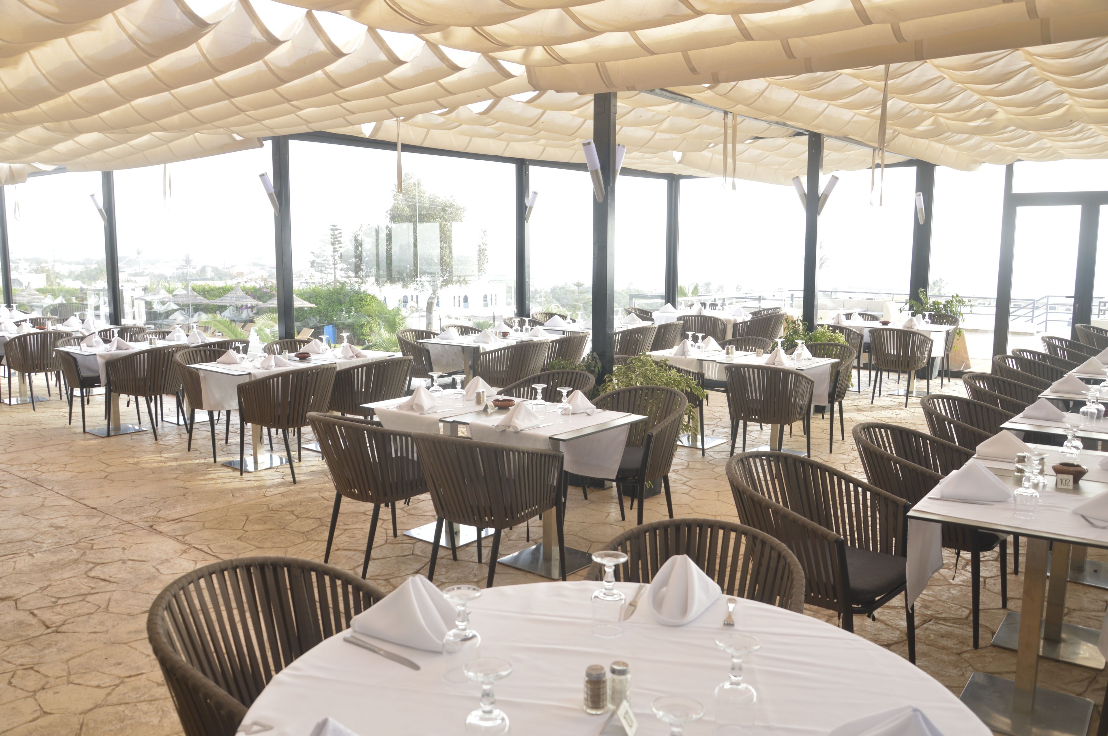 An outdoor dining area with tables and chairs, large windows, and a view of the sea.