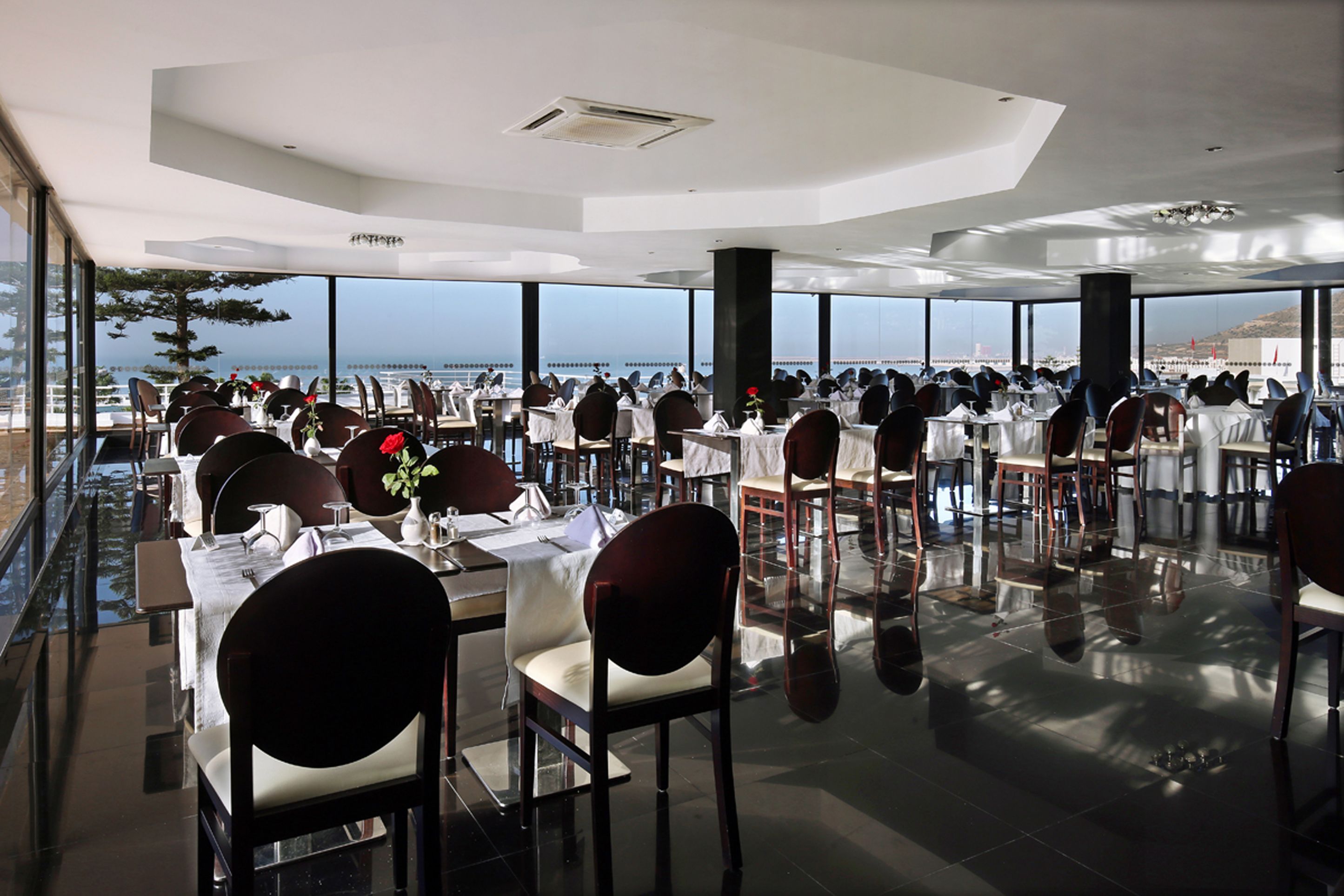 A spacious restaurant with tables and chairs arranged for dining, large windows offering a view of the sea, and a modern ceiling design.