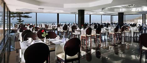 Un restaurant spacieux, avec des tables et des chaises disposées pour accueillir les clients, de grandes fenêtres offrant une vue sur la mer et un design moderne au niveau du plafond.