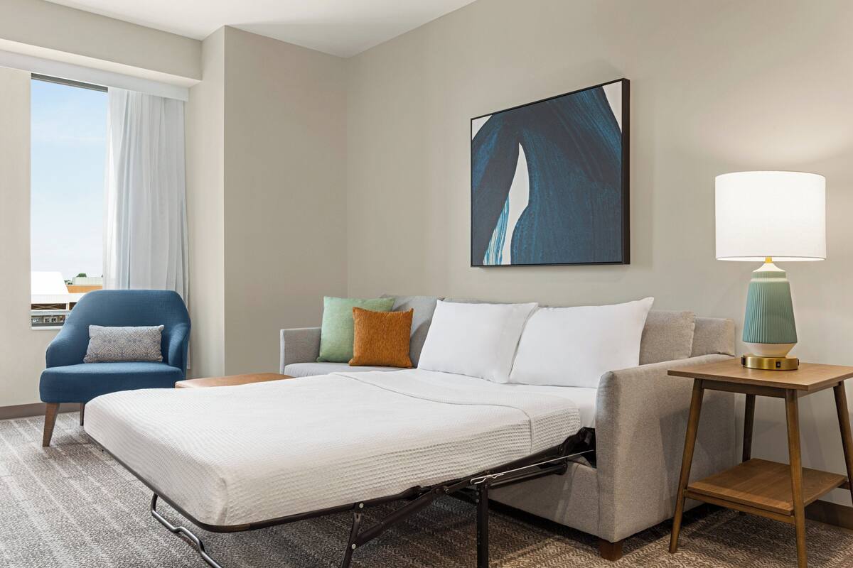 Una habitación de hotel con una cama, una silla, un sofá, una lámpara y un cuadro en la pared.
