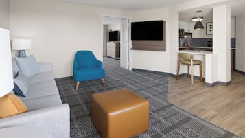 A modern hotel room with a grey sofa, a blue armchair, a brown ottoman, a flat-screen TV, a kitchenette, and a patterned rug.