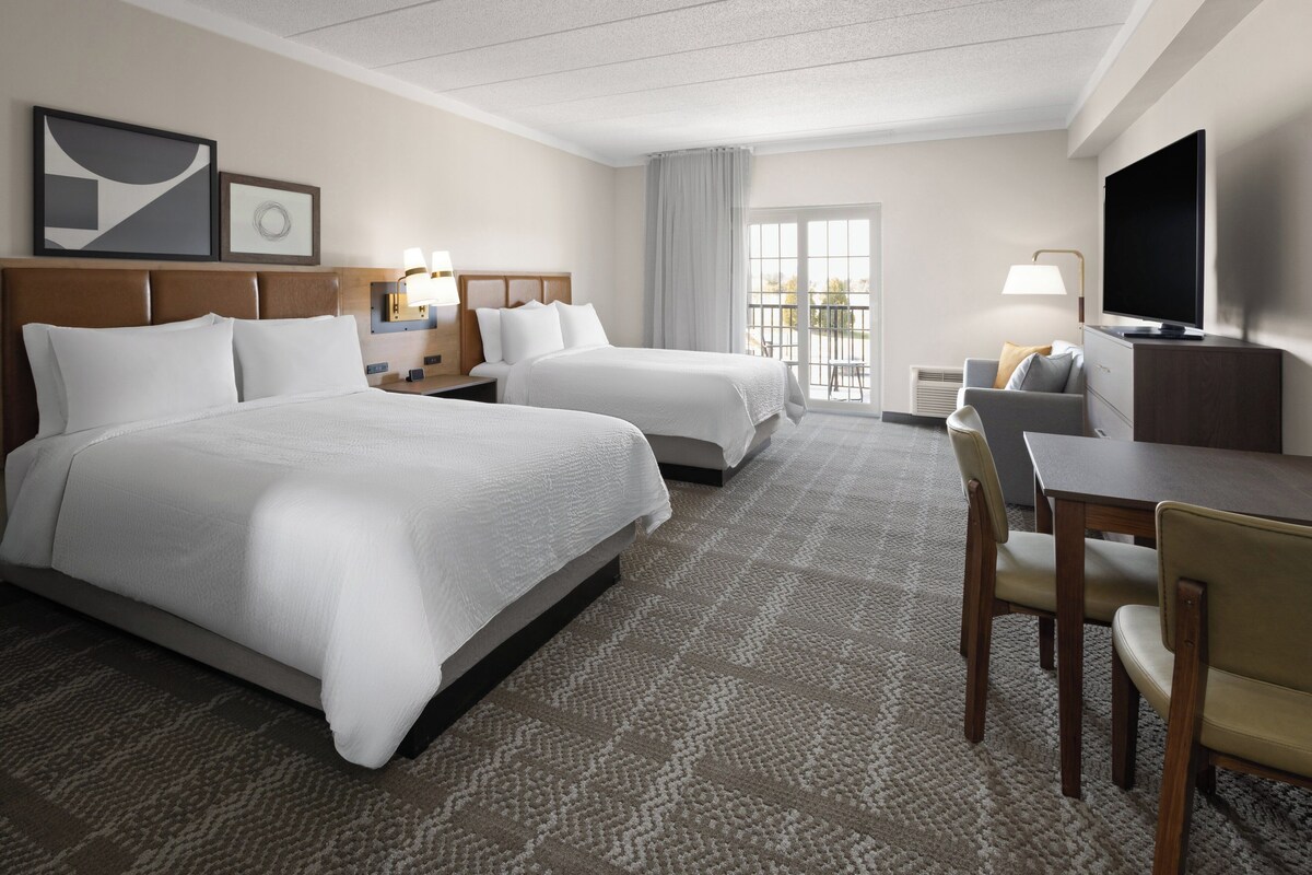 A hotel room with two beds, a desk, a chair, a TV, and a balcony with a view.