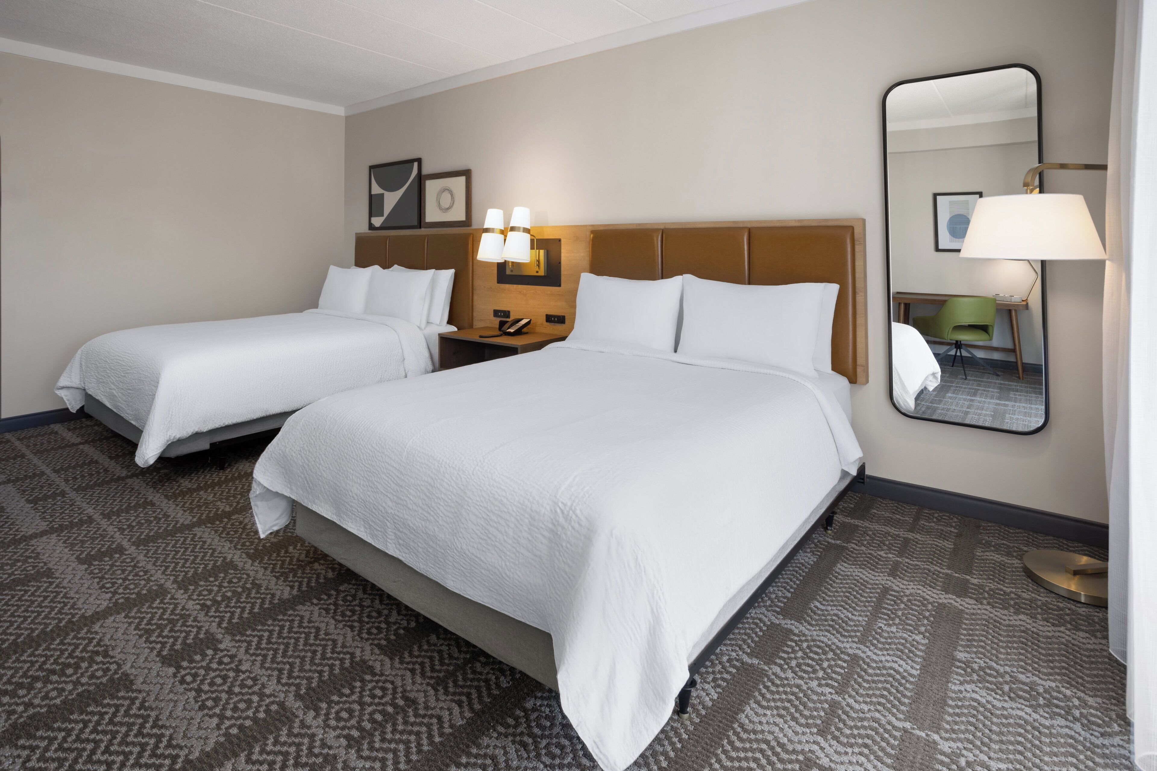 Une chambre d’hôtel avec deux lits, une table de chevet avec une lampe, un bureau avec une chaise et un grand miroir.