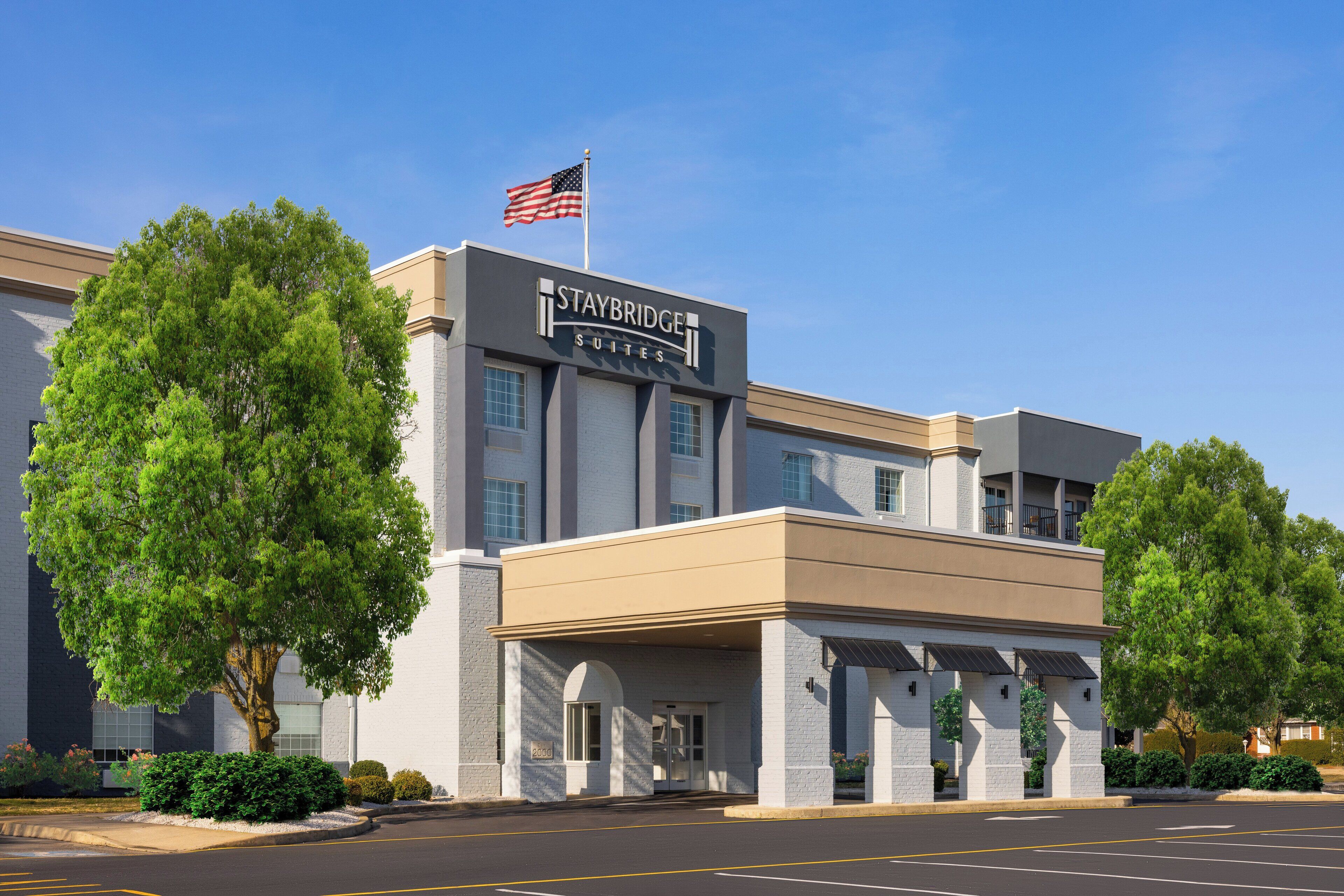 A Staybridge Suites hotel with a clear entrance, American flag, and a parking lot.