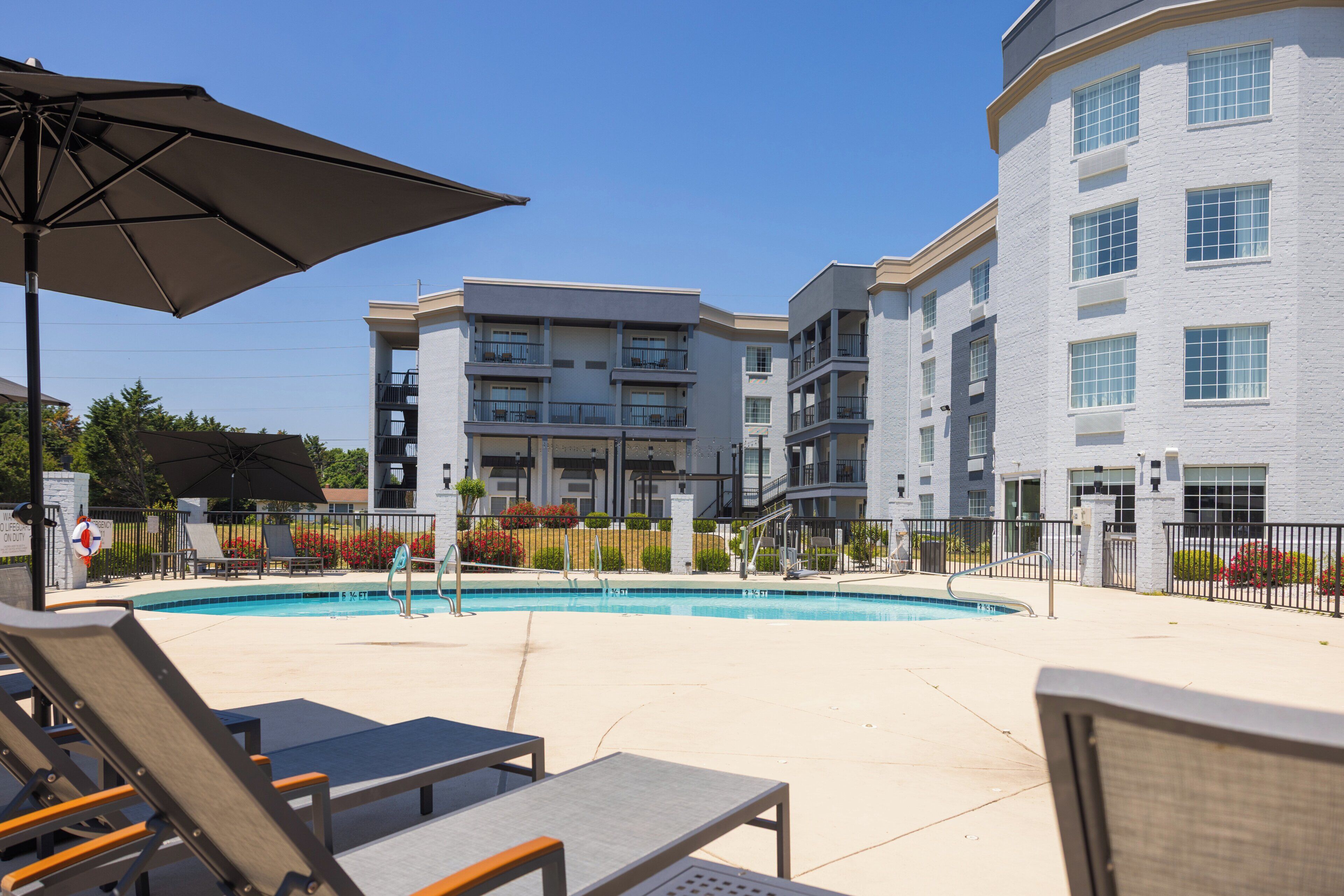 A pool area with lounge chairs and umbrellas, a building with multiple balconies, and a clear blue sky.