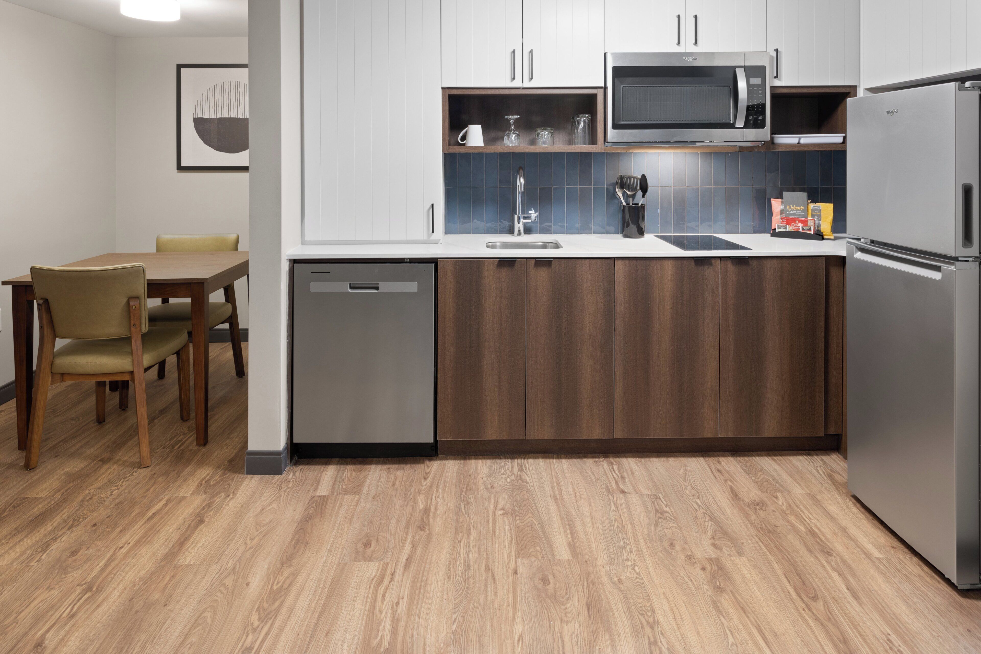 A modern kitchen with a dishwasher, microwave, and refrigerator. There is a dining table with chairs and a sink with a faucet.