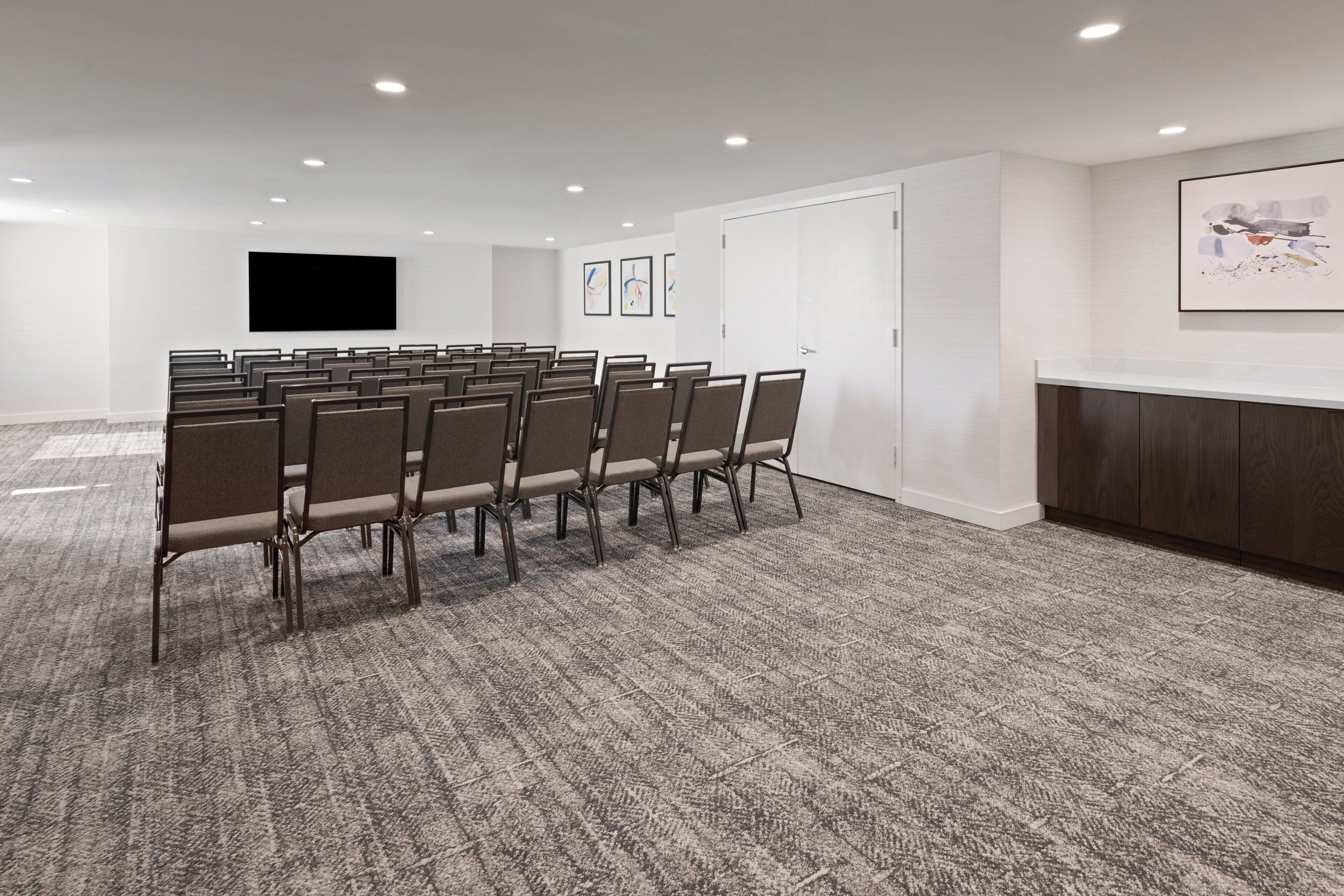 A conference room with rows of chairs, a large flat-screen TV, framed artwork on the walls, and a wooden cabinet.