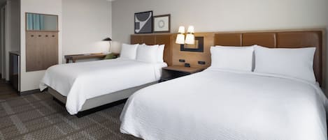 A hotel room with two beds, a desk, a lamp, and a wall-mounted light fixture.