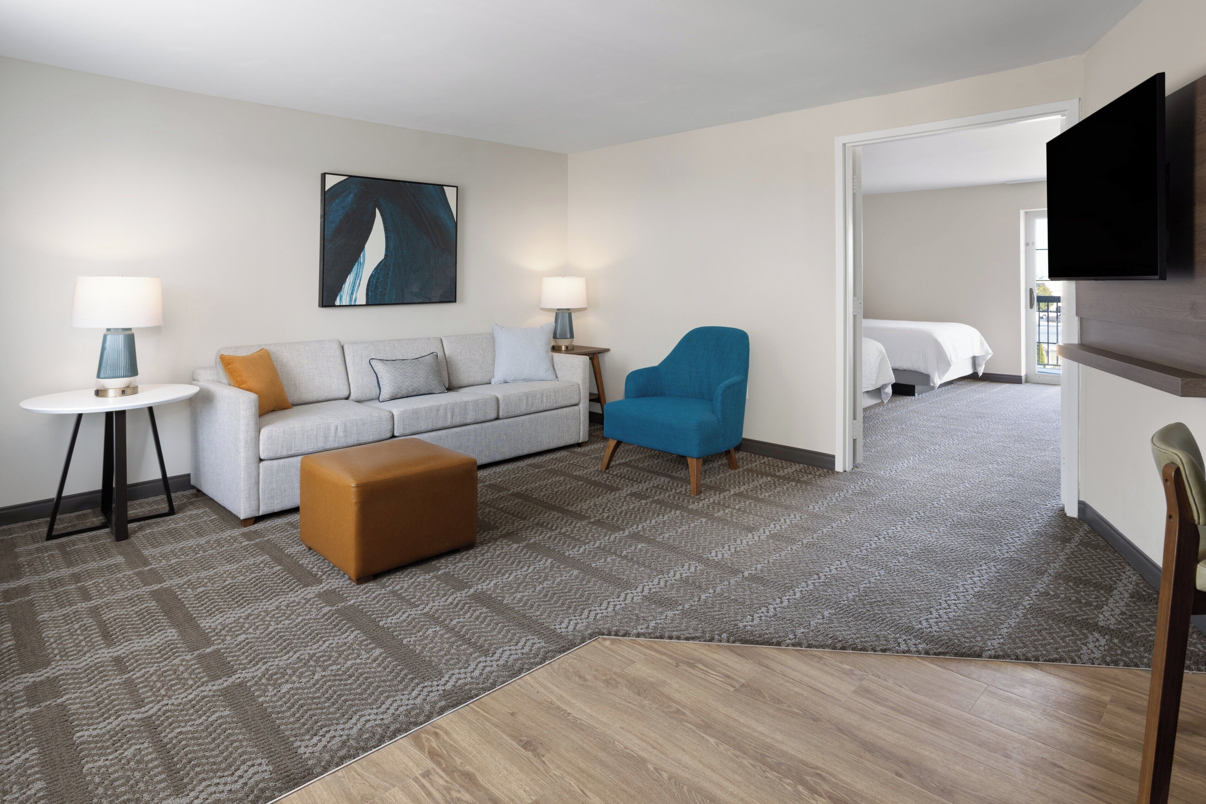 A modern hotel room with a grey sofa, a blue armchair, a brown ottoman, a small round table with a lamp, and a flat-screen TV mounted on the wall.