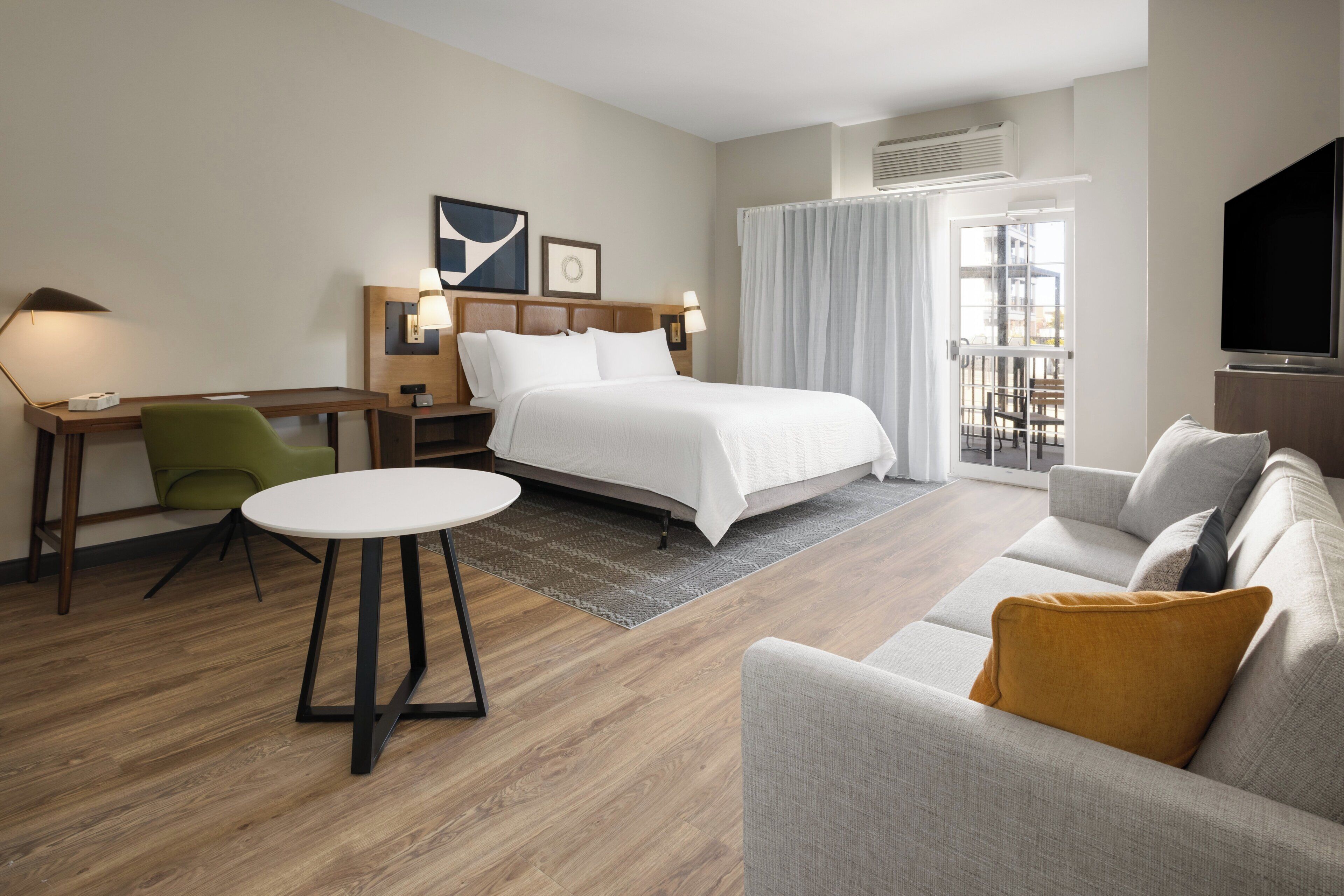 A hotel room with a bed, a sofa, a desk with a chair, a TV, and a large window.