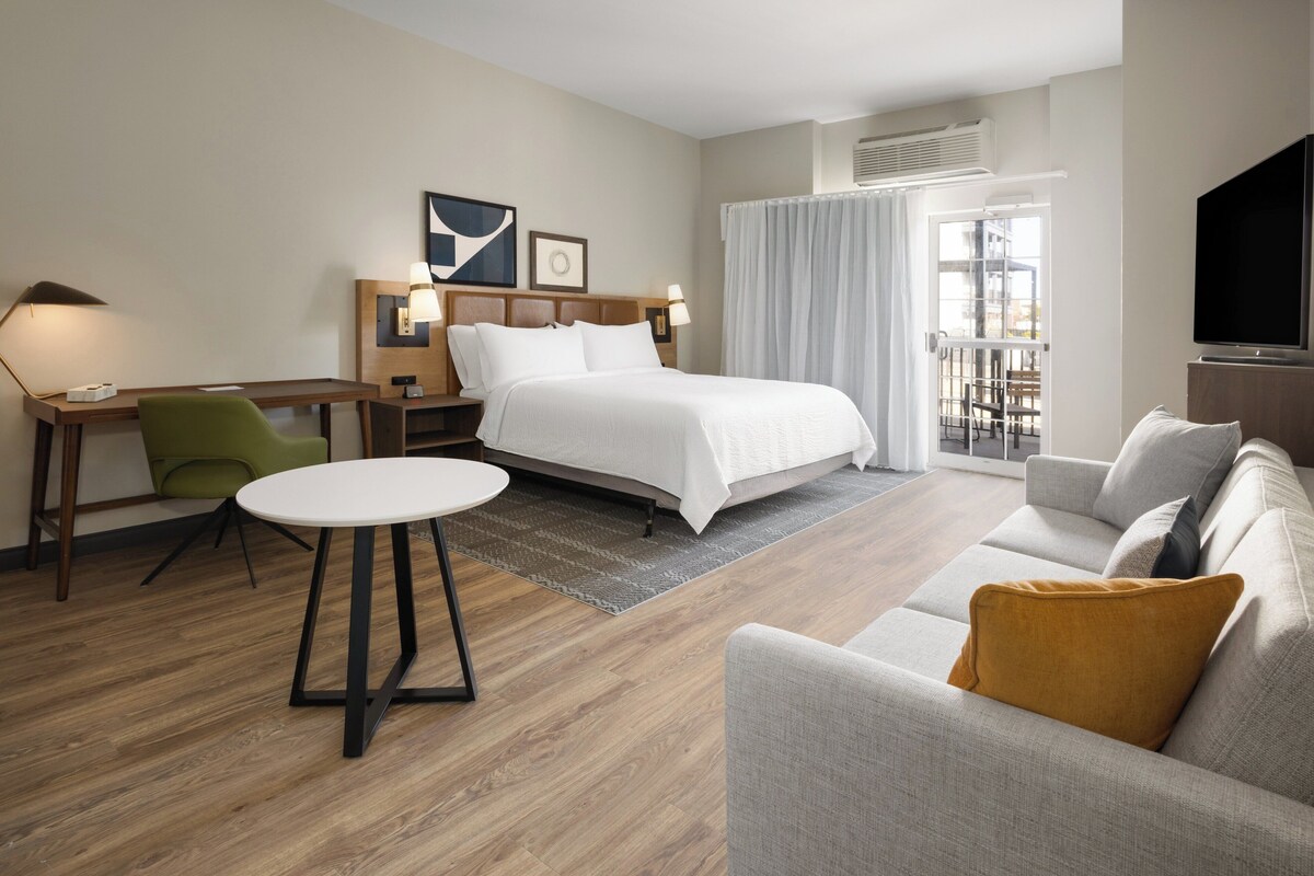 A hotel room with a bed, a sofa, a desk with a chair, a TV, and a large window.