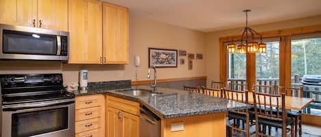 A kitchen with wooden cabinets, stainless steel appliances, a granite countertop, and a dining area with wooden chairs and a chandelier.