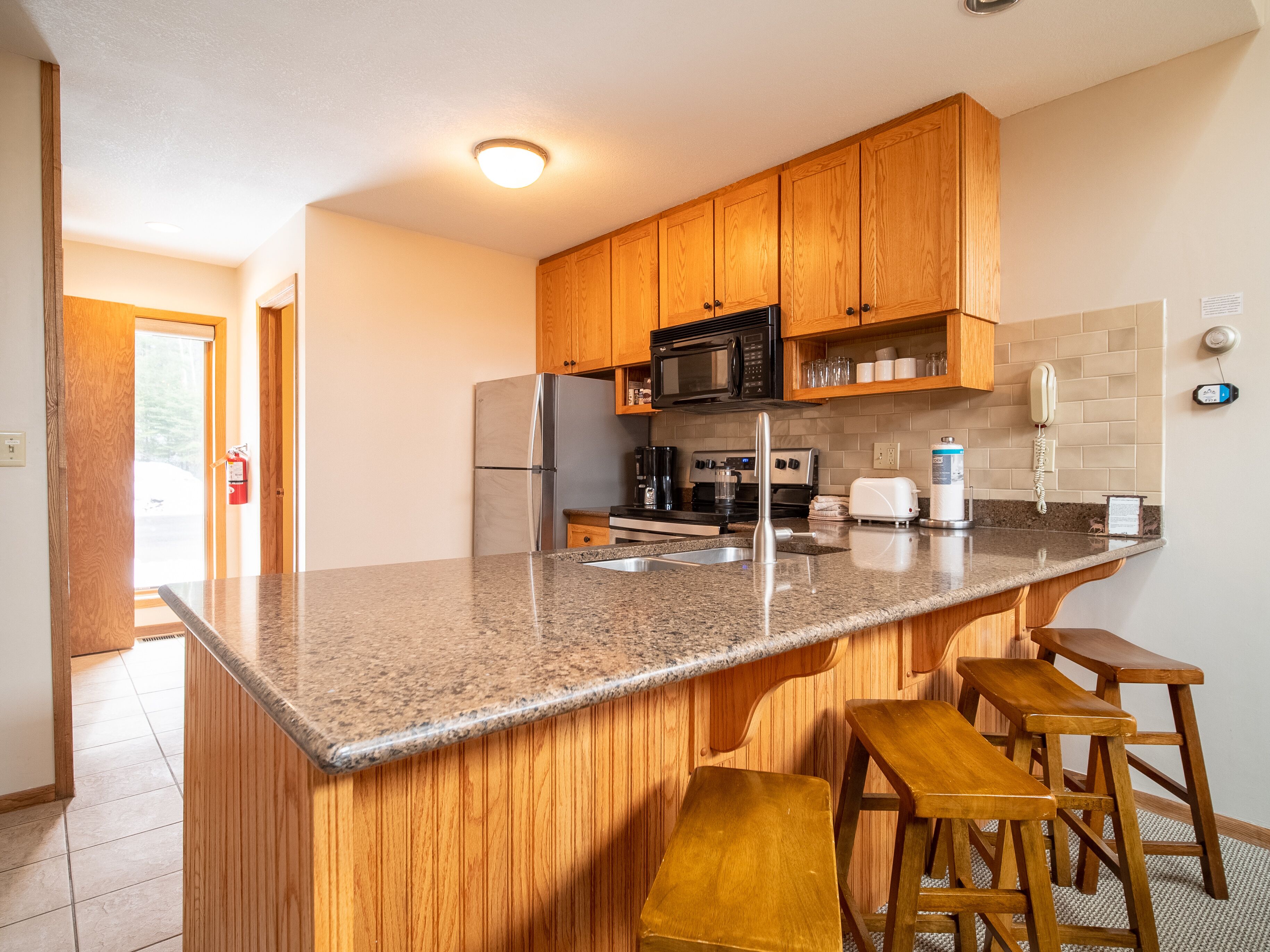 A kitchen with wooden cabinets, granite countertops, a stainless steel refrigerator, a microwave, a stove, and a sink. There are three wooden stools and a fire extinguisher near the door.