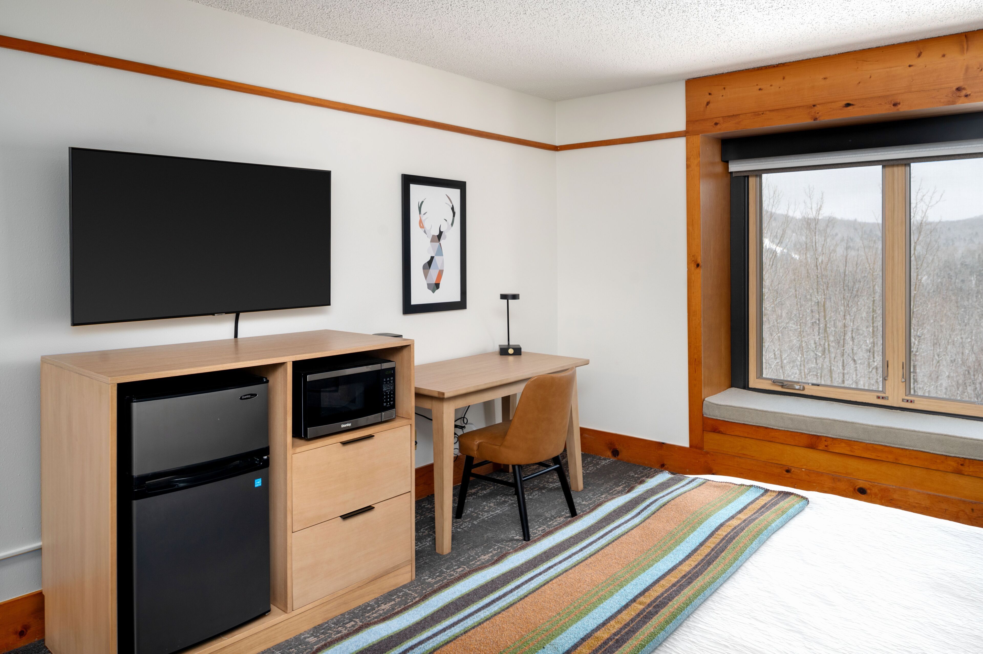 A modern hotel room with a bed, a desk, a flat-screen TV, a refrigerator, and a window with a view of trees.
