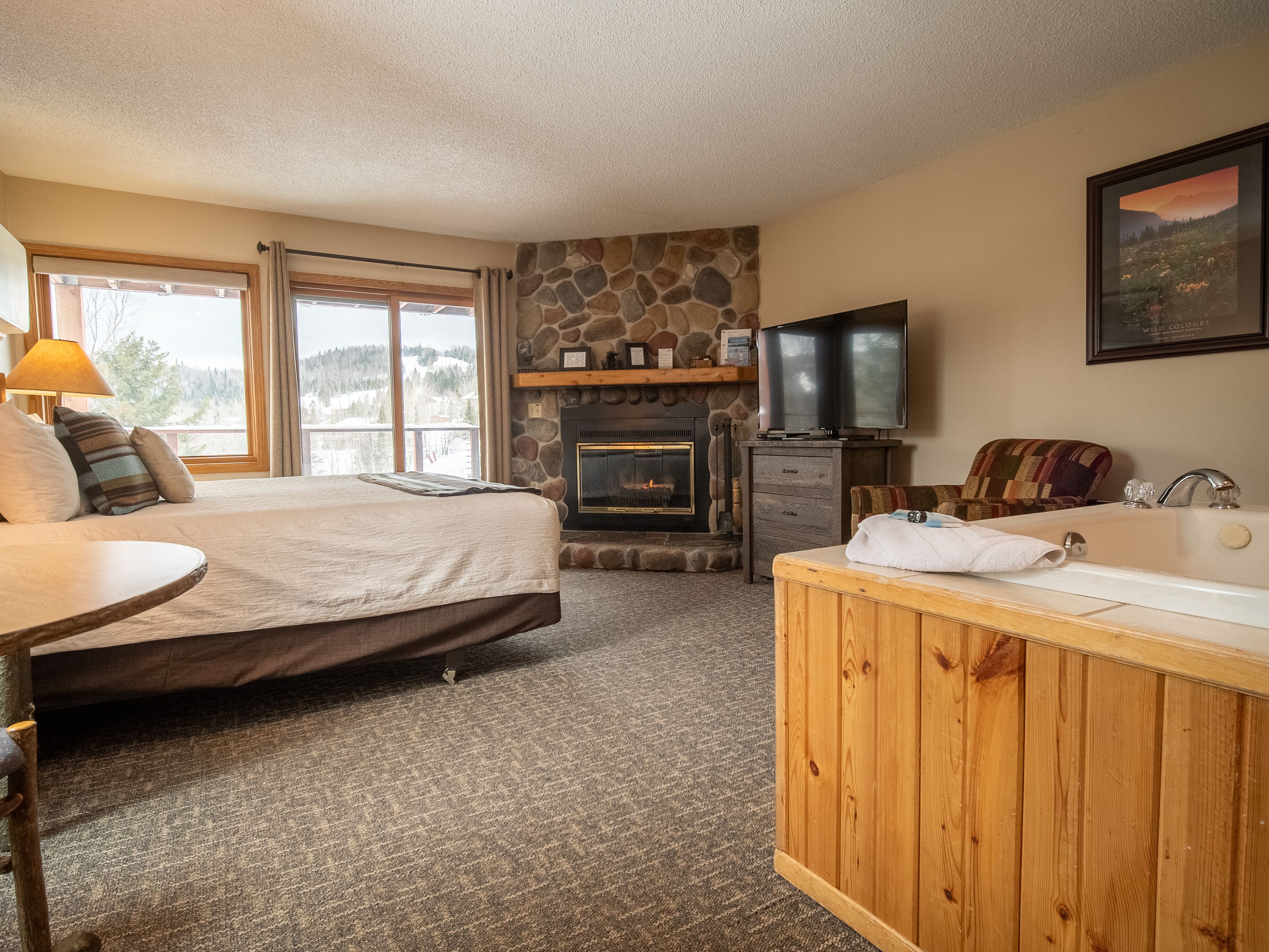 Une chambre à coucher comprenant un lit, une cheminée en pierre, un téléviseur, une coiffeuse en bois avec un lavabo et une fenêtre donnant sur un paysage enneigé.