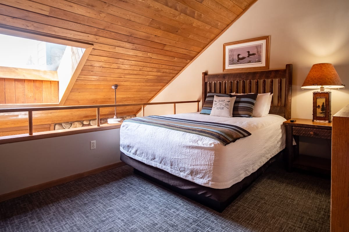 Une chambre avec un plafond en pente, un lit avec une literie à rayures, une table de chevet avec une lampe et un tableau encadré au mur.