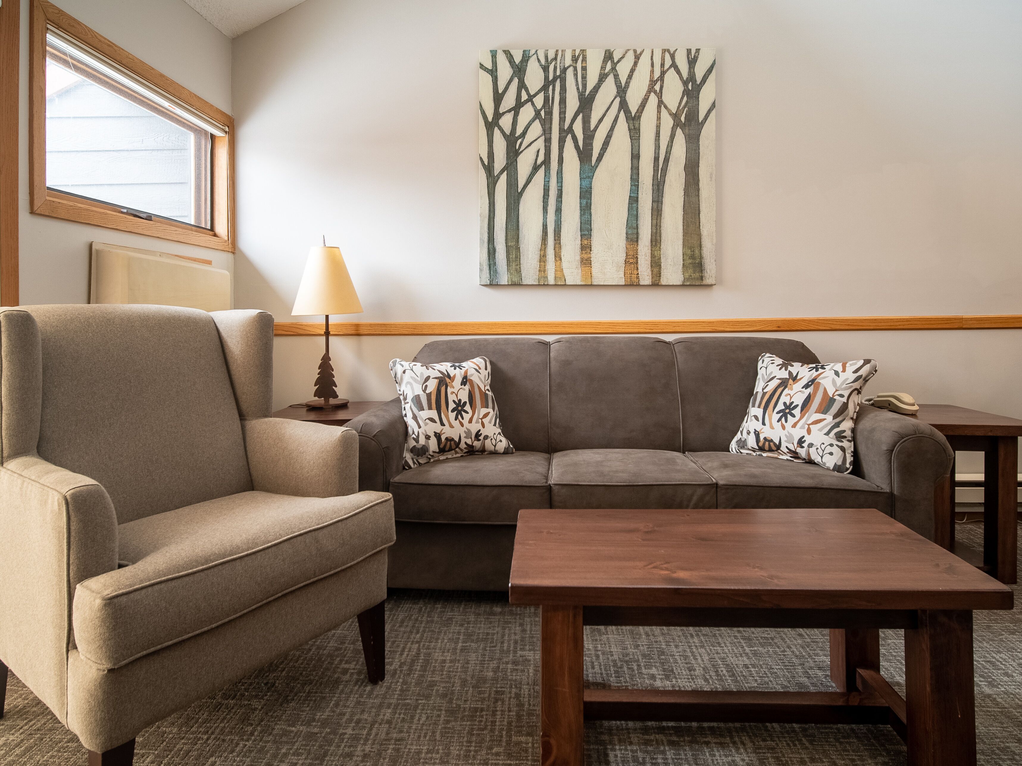A living room with a sofa, armchair, and a wooden coffee table. There is a lamp on a side table, a painting on the wall, and a window with blinds.