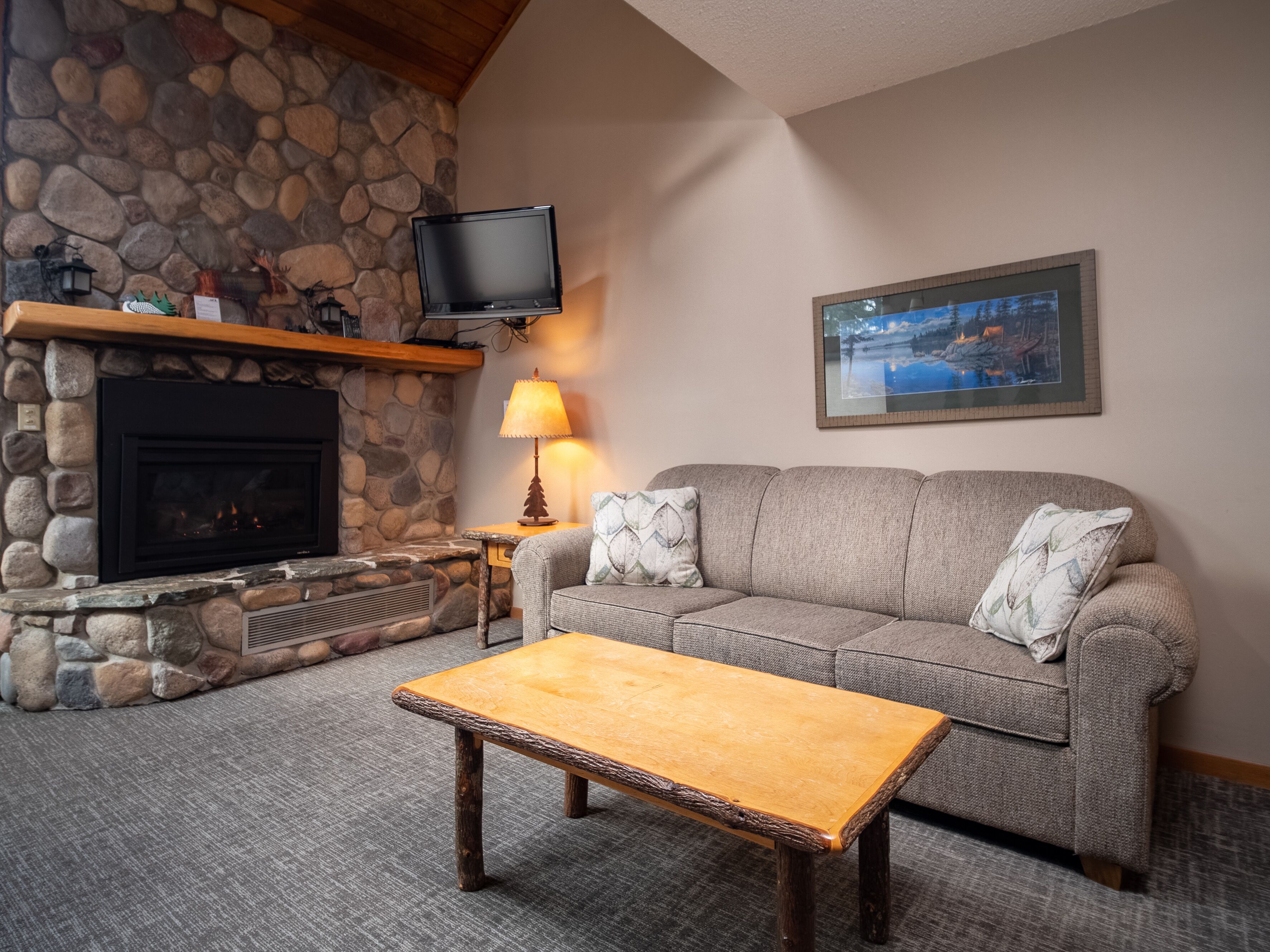A cozy living room with a stone fireplace, a flat-screen TV mounted above it, a gray sofa with patterned cushions, and a wooden coffee table.