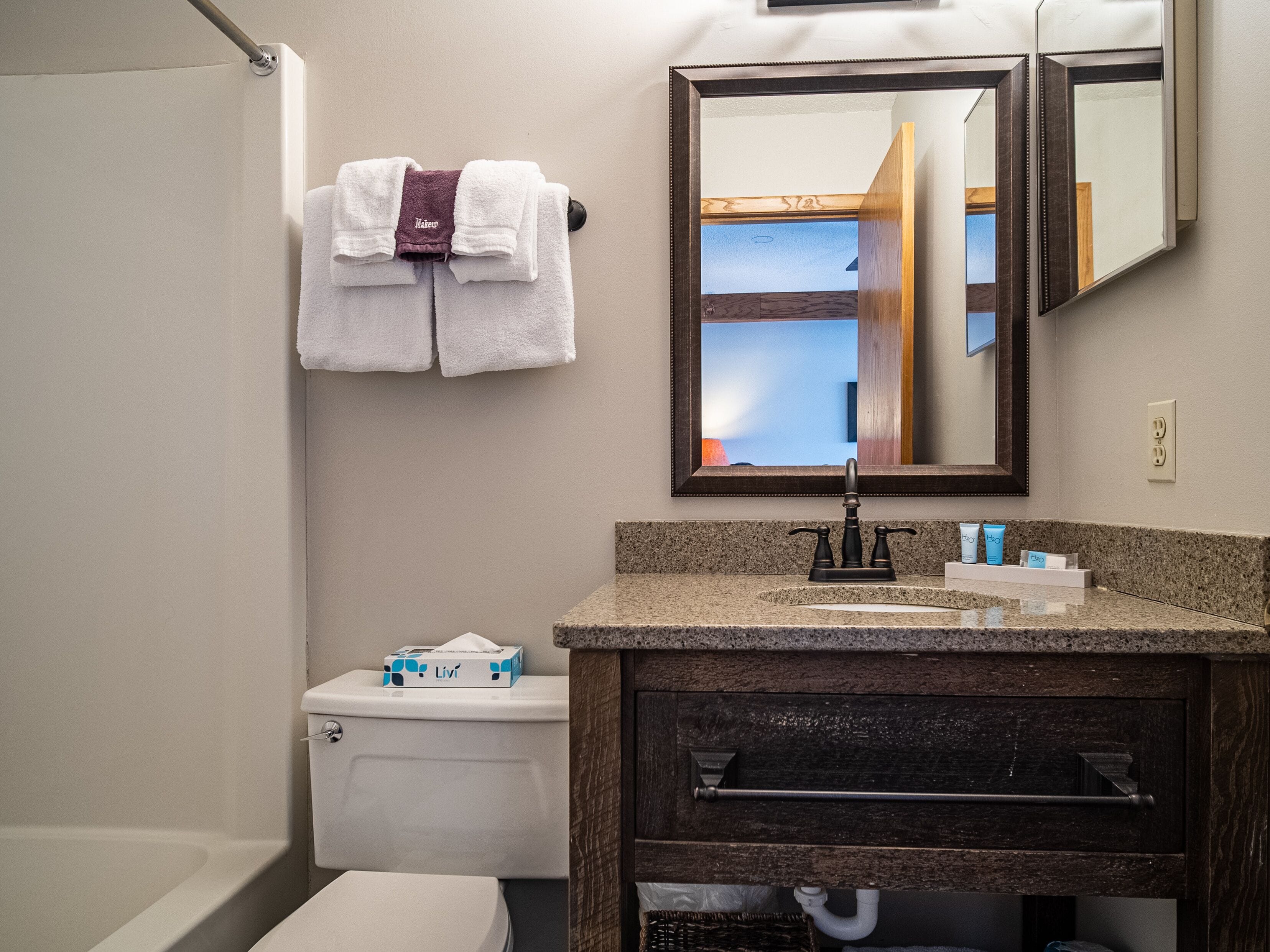 A bathroom with a toilet, sink, and mirror. There are towels hanging on a rack and a box of tissues on the toilet.
