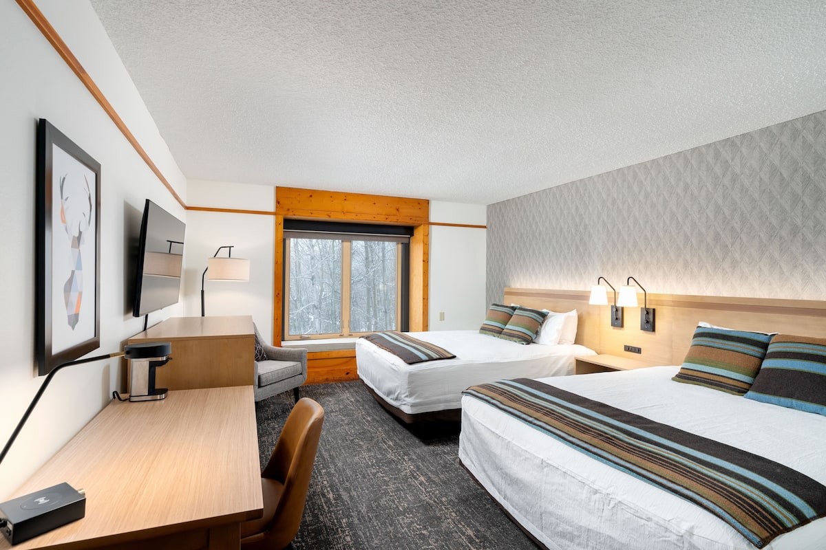 Une chambre d’hôtel avec deux lits, un bureau, une chaise, une télévision et une fenêtre donnant sur des arbres.