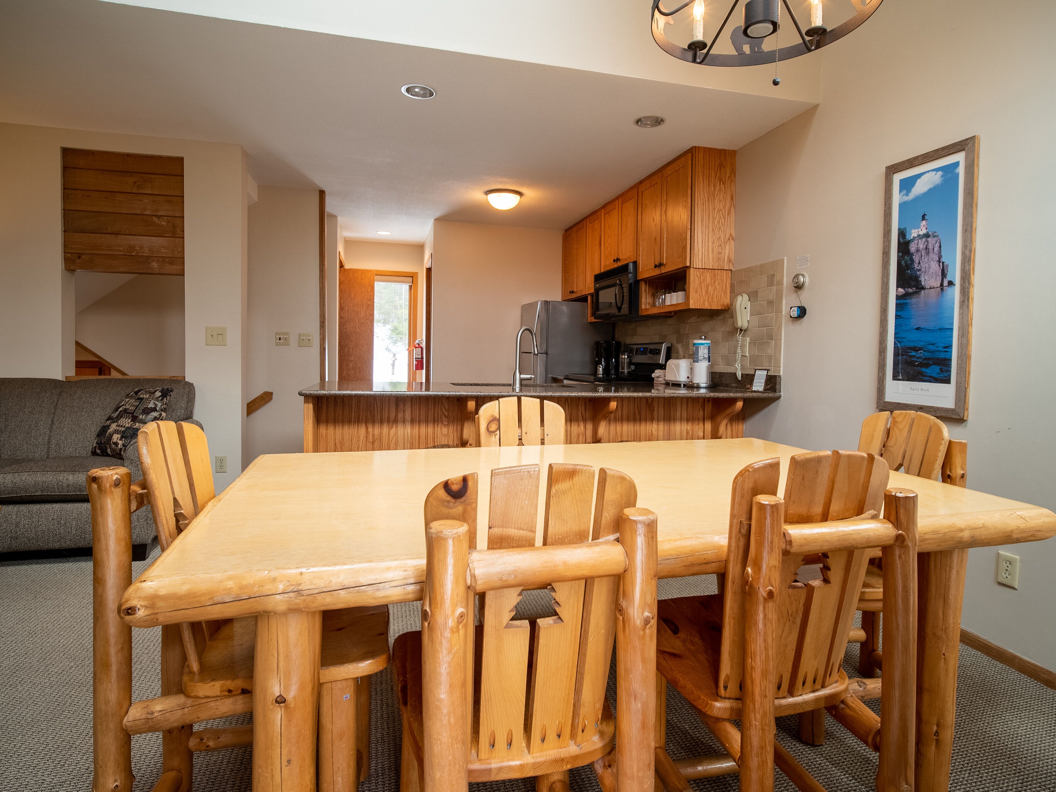 A wooden dining table with six chairs, a kitchen with wooden cabinets, a refrigerator, and a microwave.