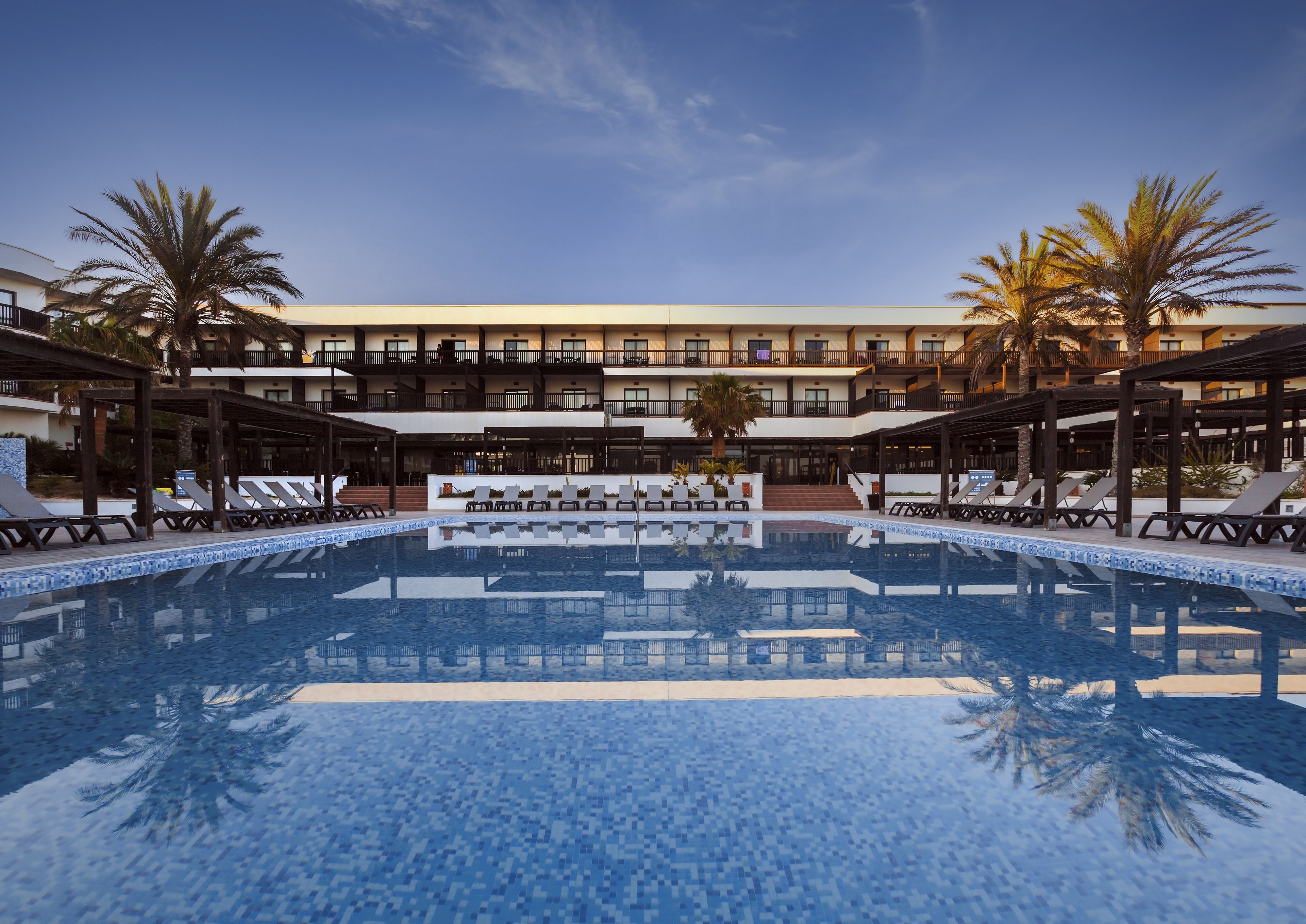 A resort pool with lounge chairs and palm trees, surrounded by a multi-story building with balconies.