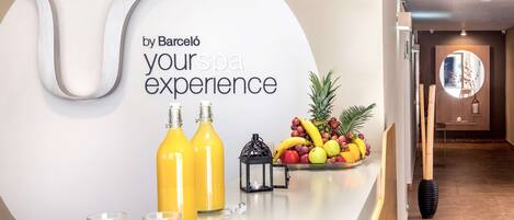 A reception area with a sign reading "Barceló yourspa experience," a fruit bowl, and two bottles of orange juice on a counter.