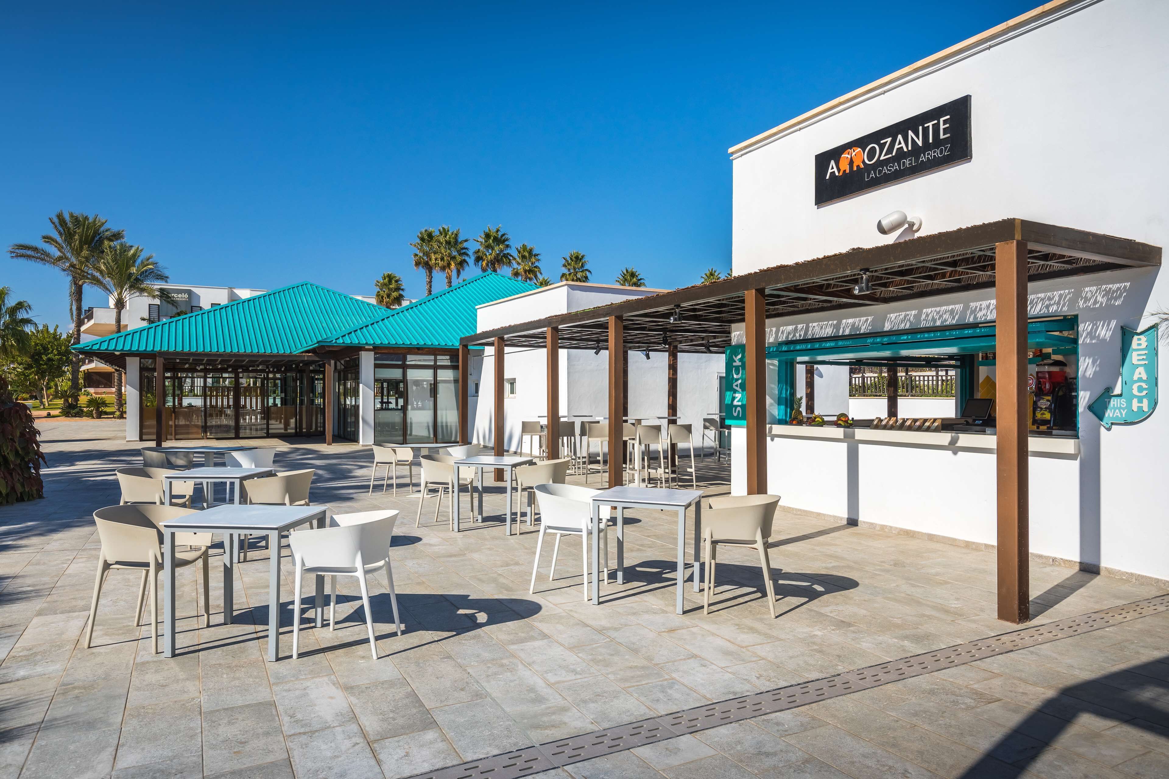 An outdoor patio area with tables and chairs, a covered seating area, and a building with a sign reading "Aroztante La Casa del Arroz".