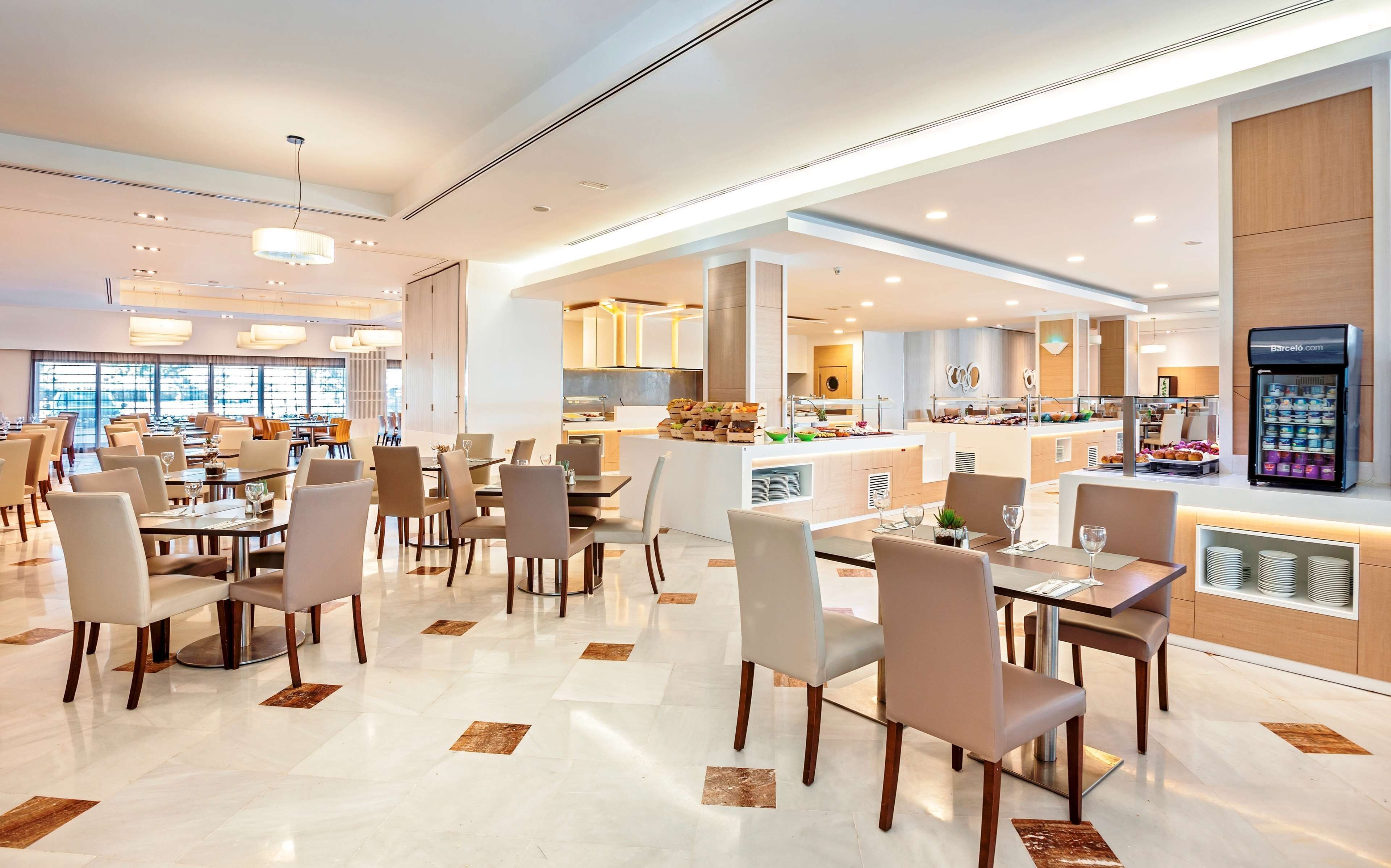 A modern hotel dining area with multiple tables and chairs, a central island with food displays, and a refrigerator stocked with beverages.