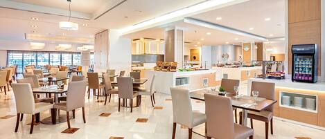 A modern hotel dining area with multiple tables and chairs, a central island with food displays, and a refrigerator stocked with beverages.