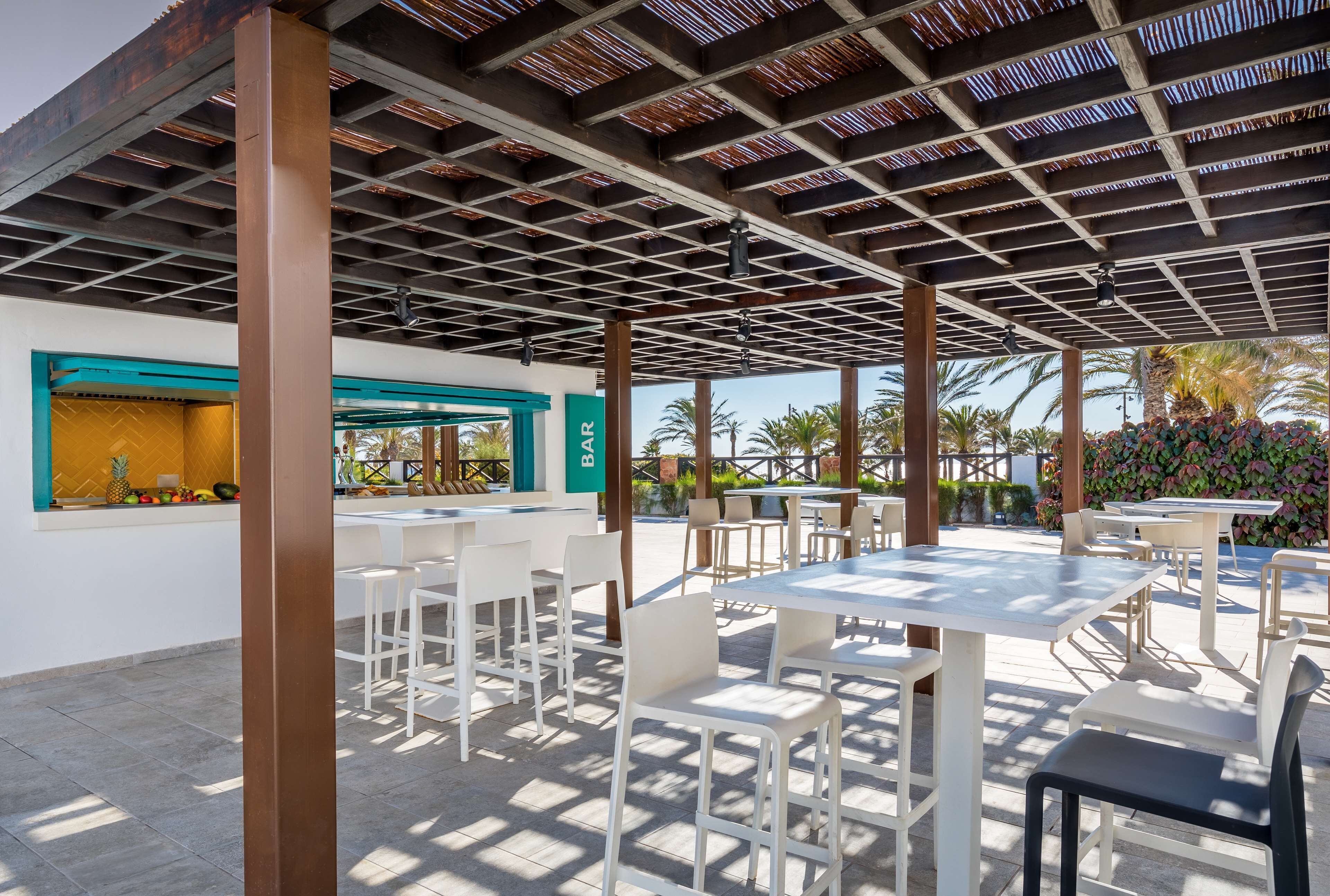 An outdoor patio with white tables and chairs, a bar area with a green accent wall, and a view of palm trees.