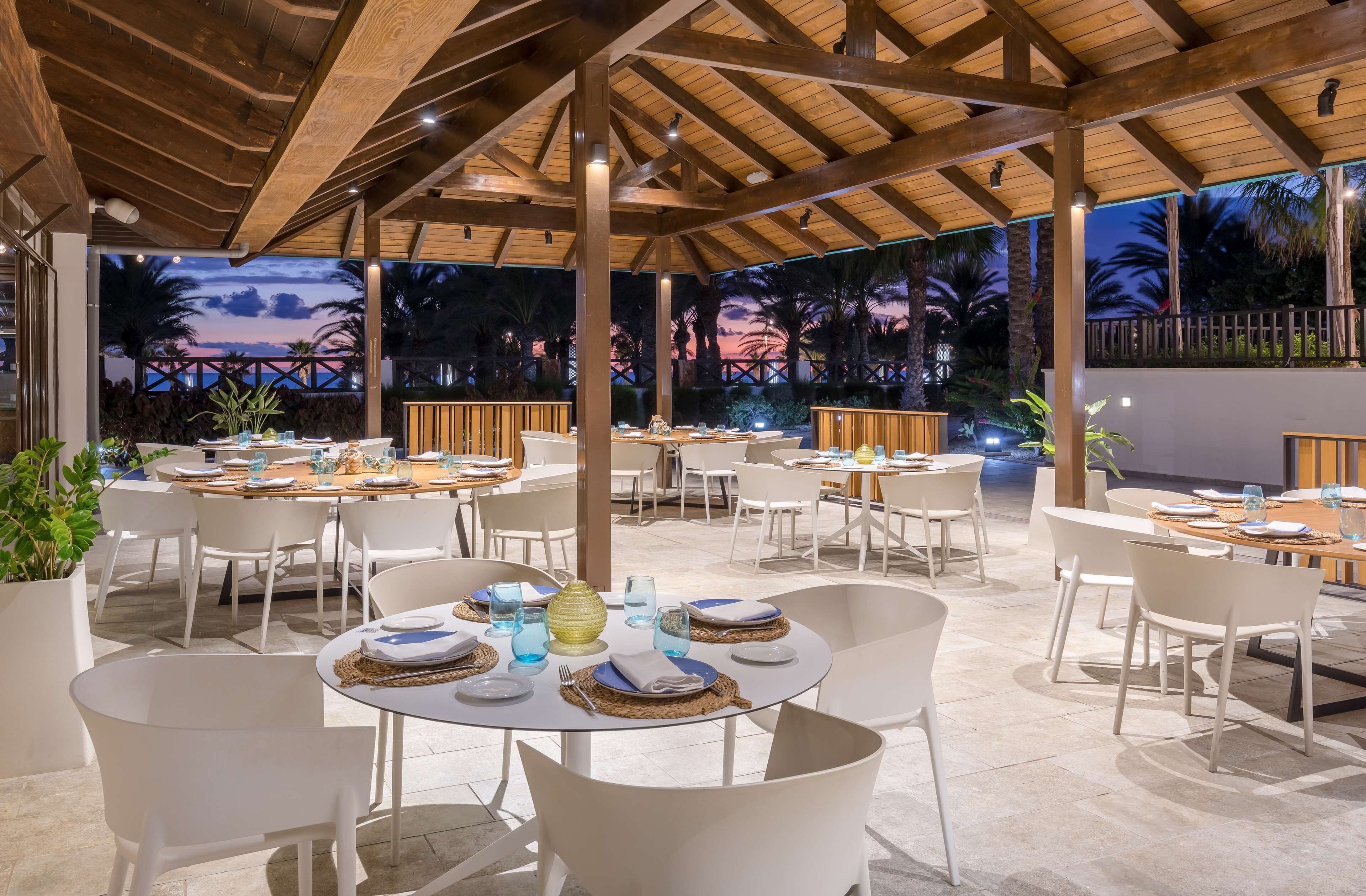 An outdoor dining area with round tables and white chairs, under a wooden pergola, with a view of palm trees and a sunset in the background.
