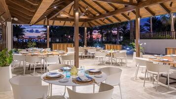 Un área de comedor al aire libre con mesas redondas y sillas blancas, bajo una pérgola de madera, con vistas a palmeras y un atardecer de fondo.