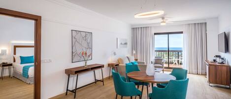 Habitación de hotel moderna con balcón, zona de comedor con sillas color turquesa, escritorio de madera y cama con almohadones azules.