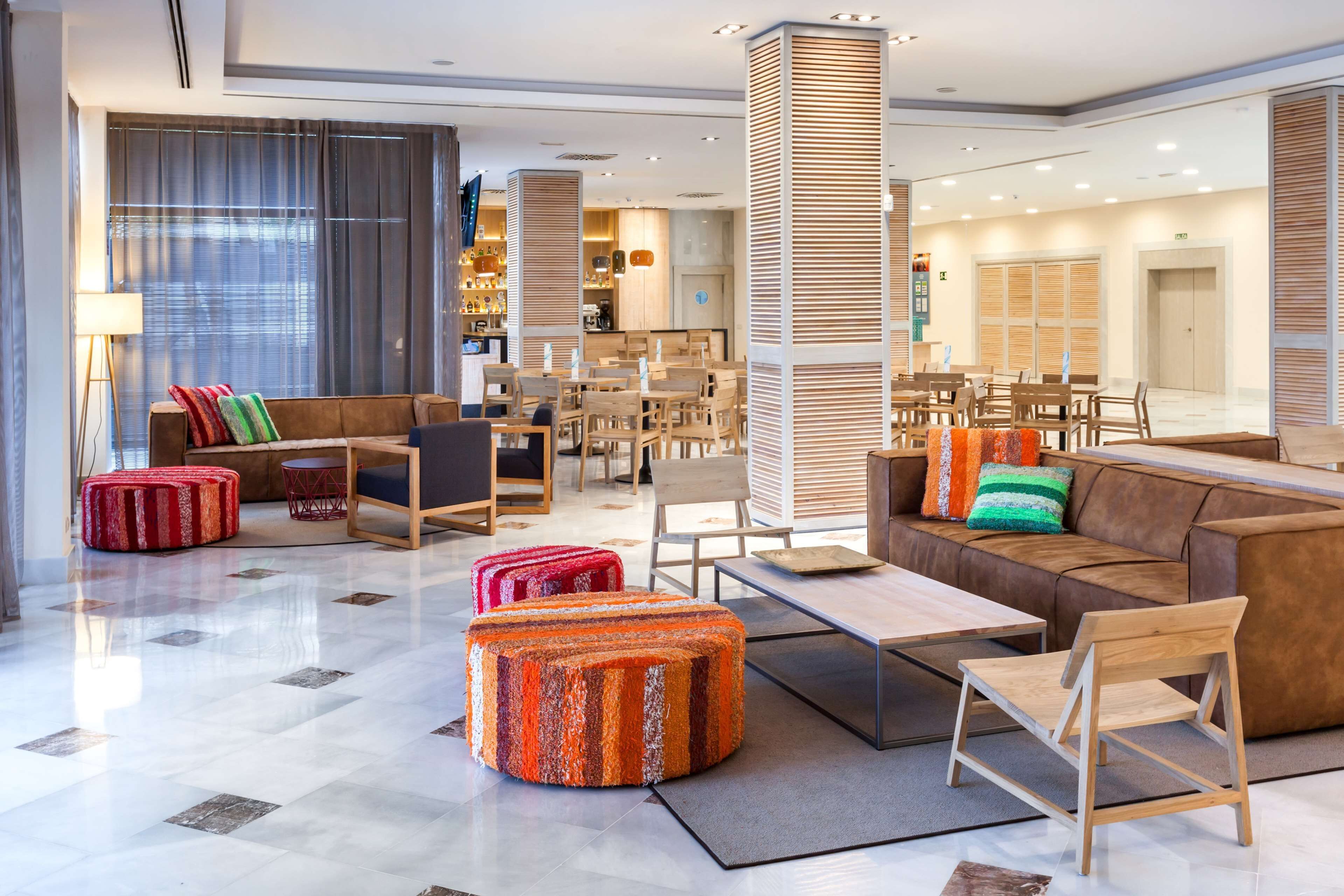 Eine großzügige Lobby mit modernem Design, ausgestattet mit einer geräumigen Sitzecke, Sofas, Poufs, einem Couchtisch und einem Holzstuhl.