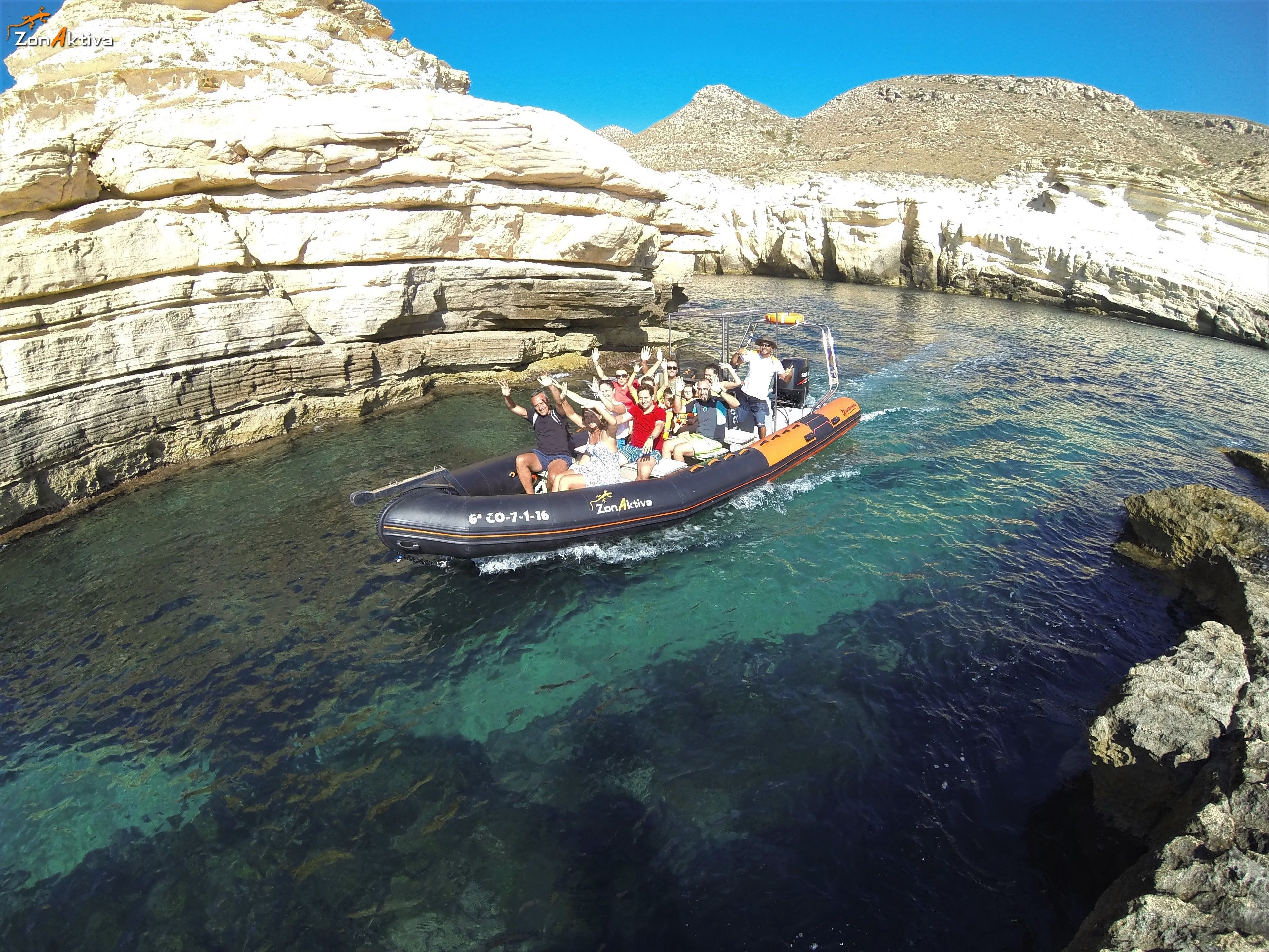 A group of people on an inflatable boat in a clear blue water body surrounded by rocky cliffs.