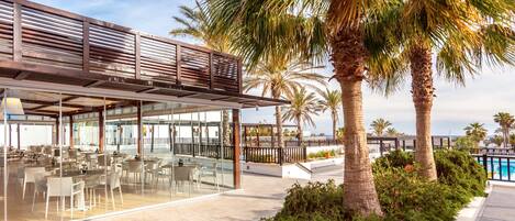 A modern outdoor dining area with glass partitions, a pool, and palm trees.