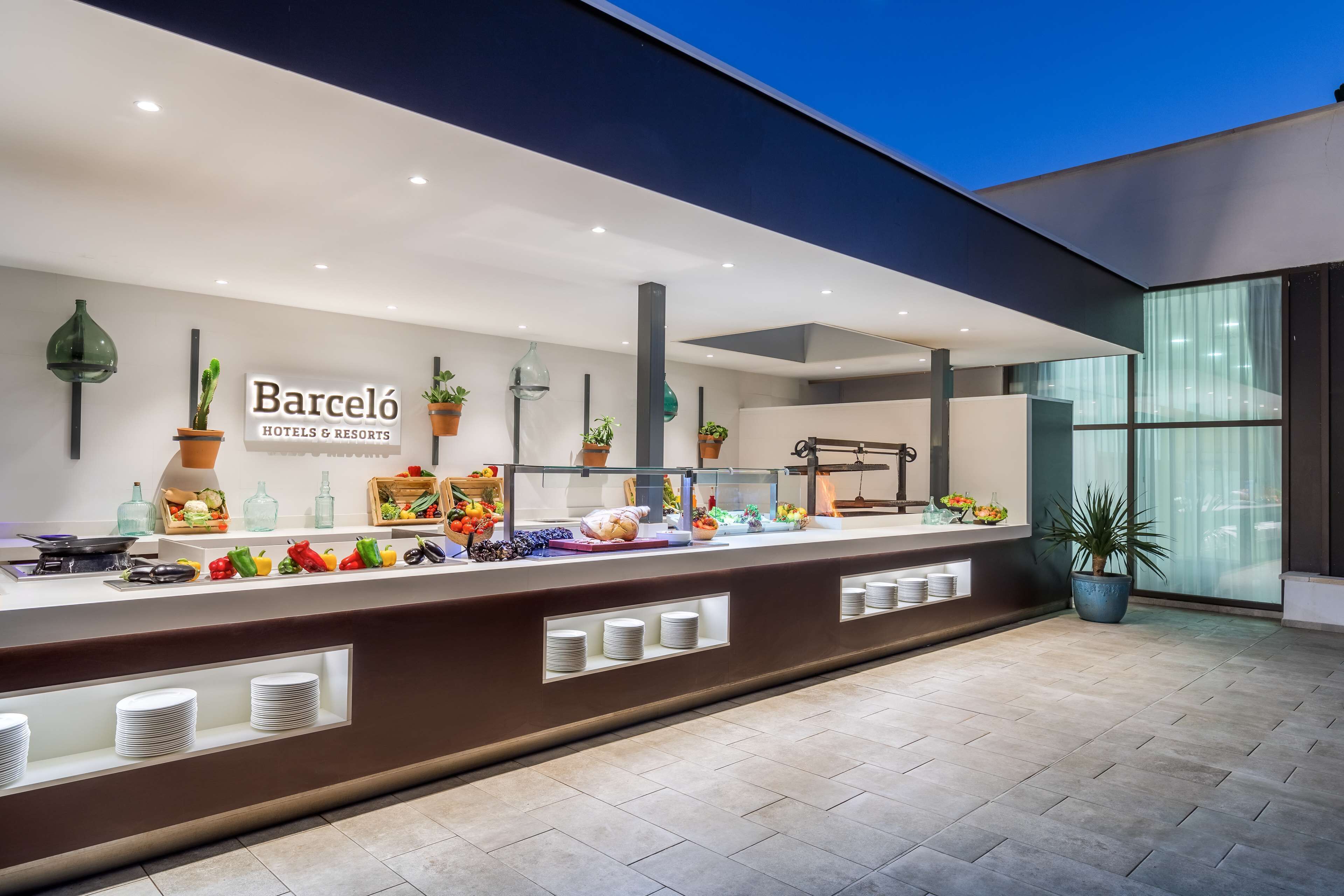 A modern hotel reception area with a buffet counter, potted plants, and a sign for Barceló Hotels & Resorts.