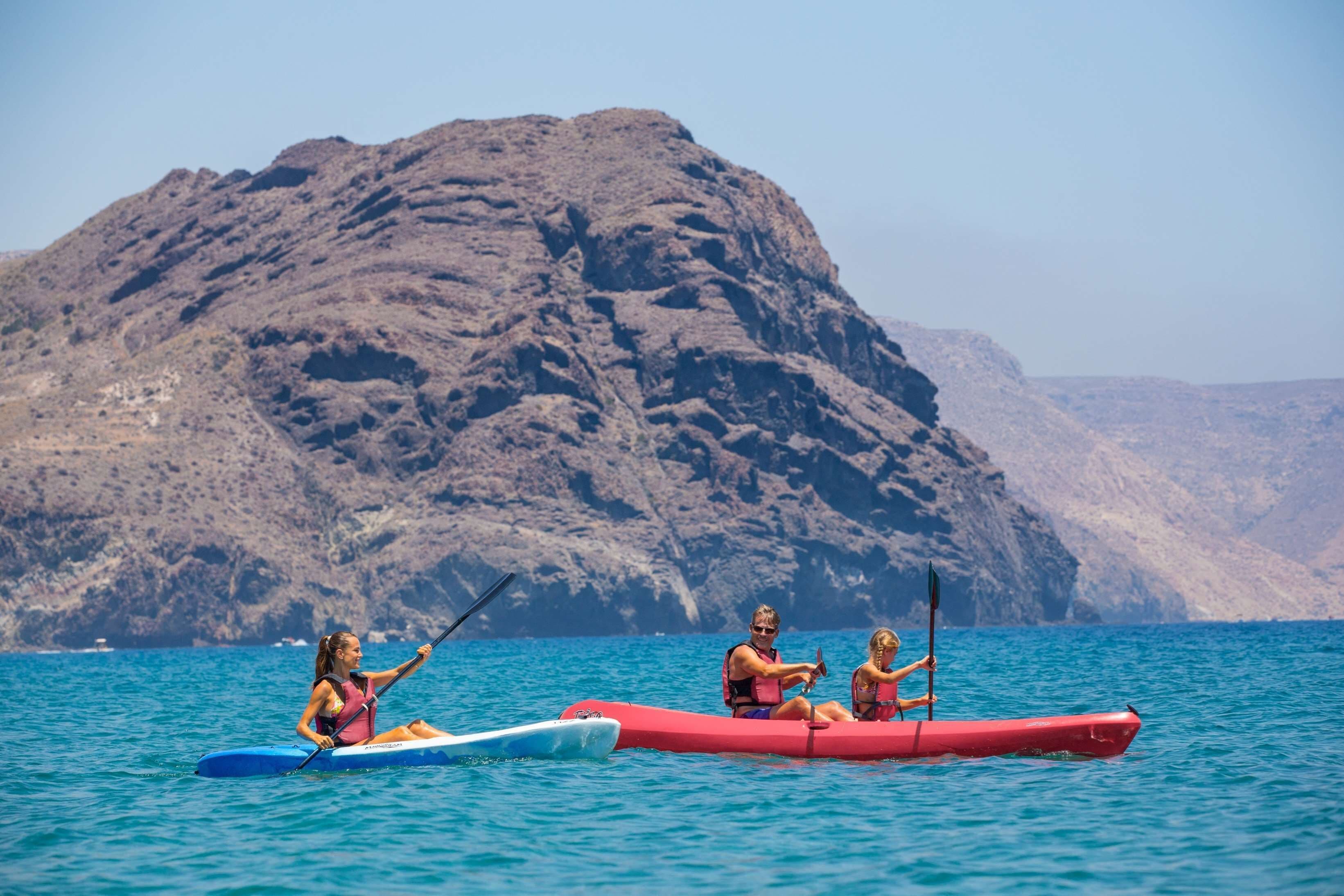 Two people kayaking on a calm sea with rocky cliffs in the background.