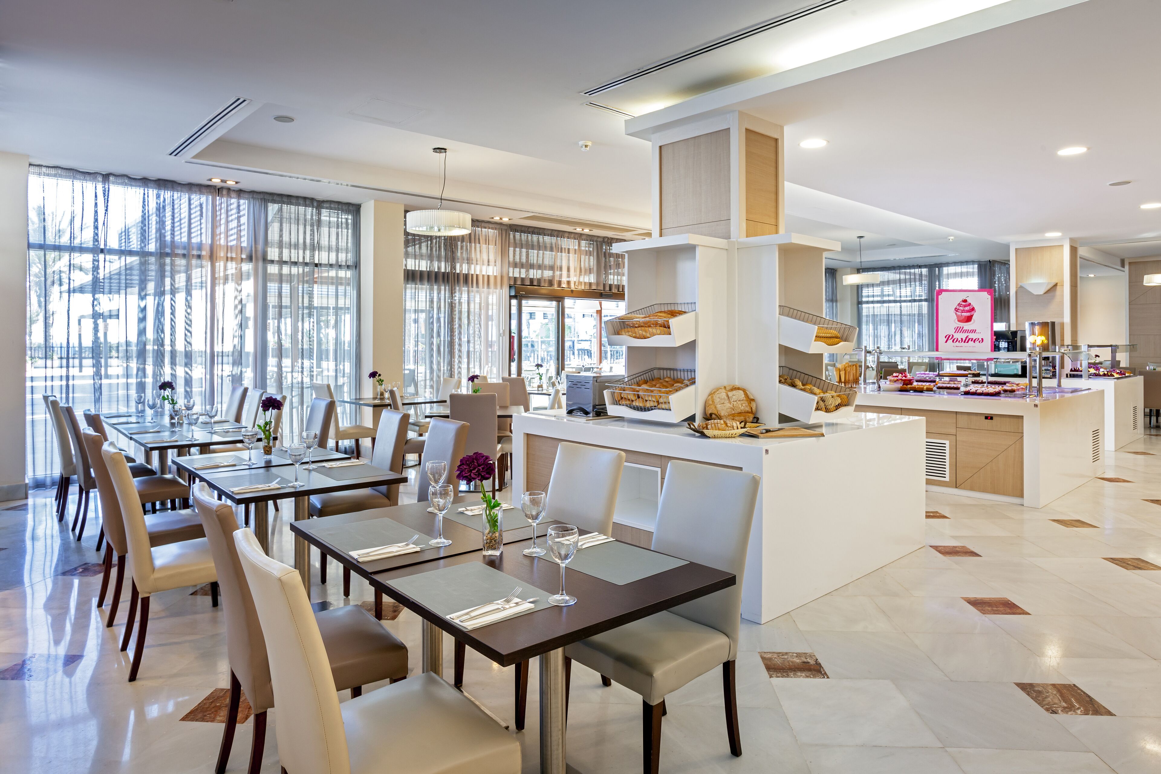 Un espace repas moderne avec des tables dressées pour le service, un comptoir central où sont exposées des pâtisseries, et une vue sur la ville grâce à de grandes fenêtres.