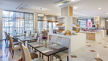 A modern dining area with tables set for service, a central counter with baked goods, and a view of the city through large windows.