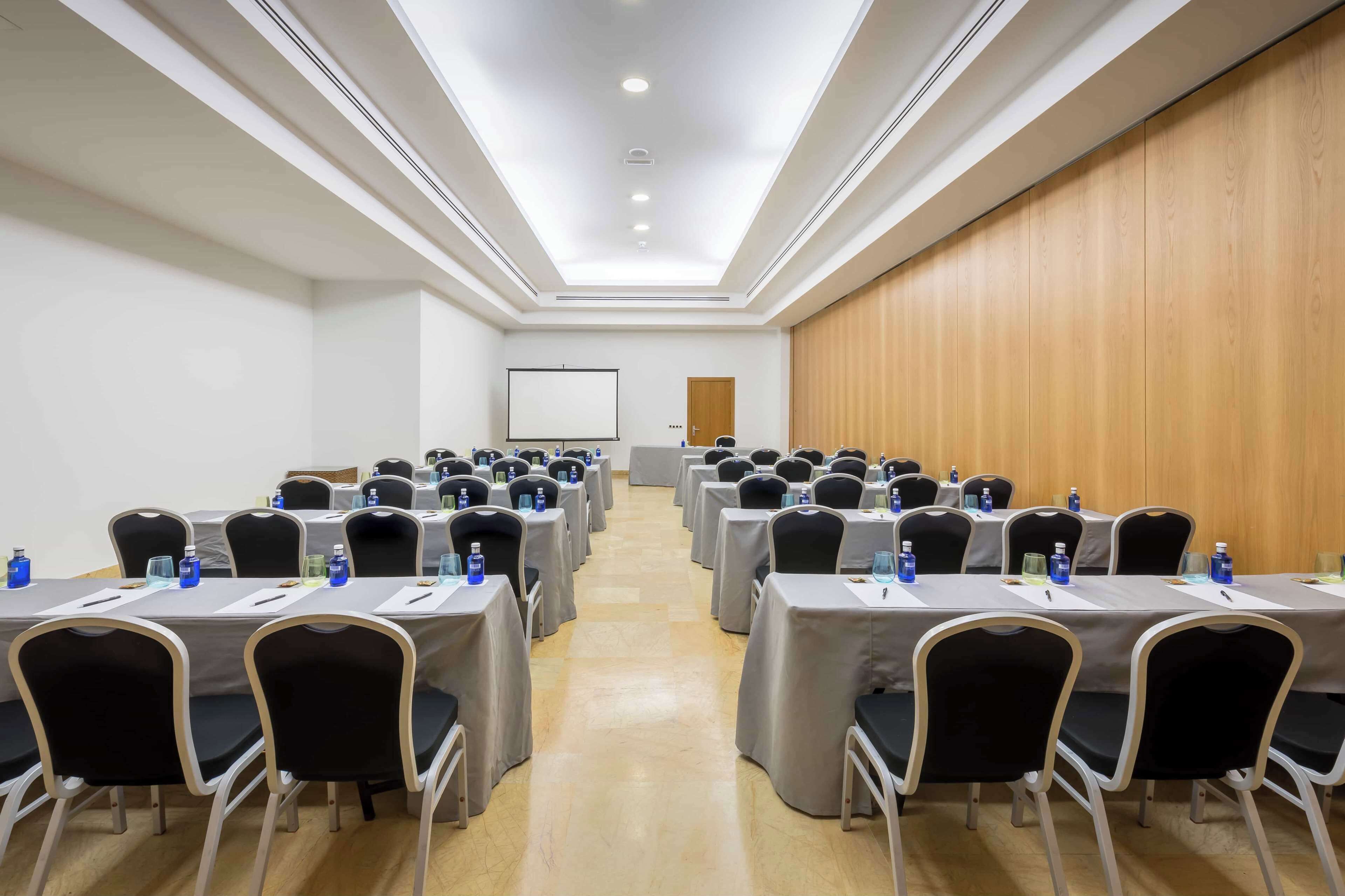 Un salón de conferencias con varias mesas y sillas, equipado con botellas de agua y tarjetas de identificación, y una pantalla de proyector al frente.