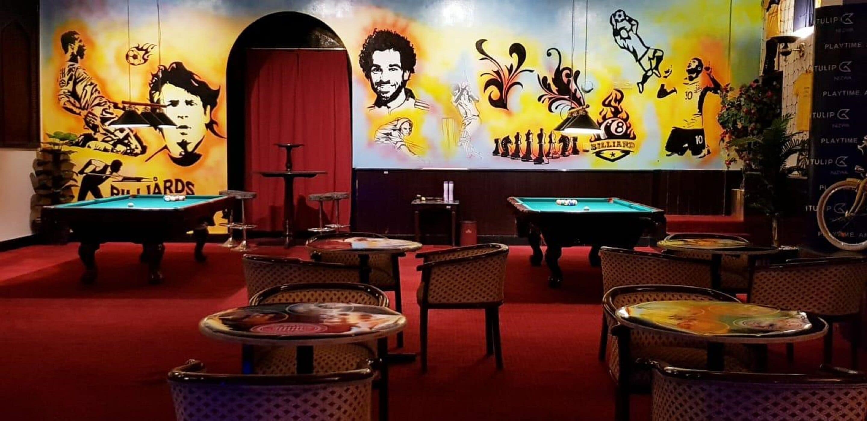 A billiards room with multiple tables and chairs, a red carpet, and a mural with billiards-related artwork.