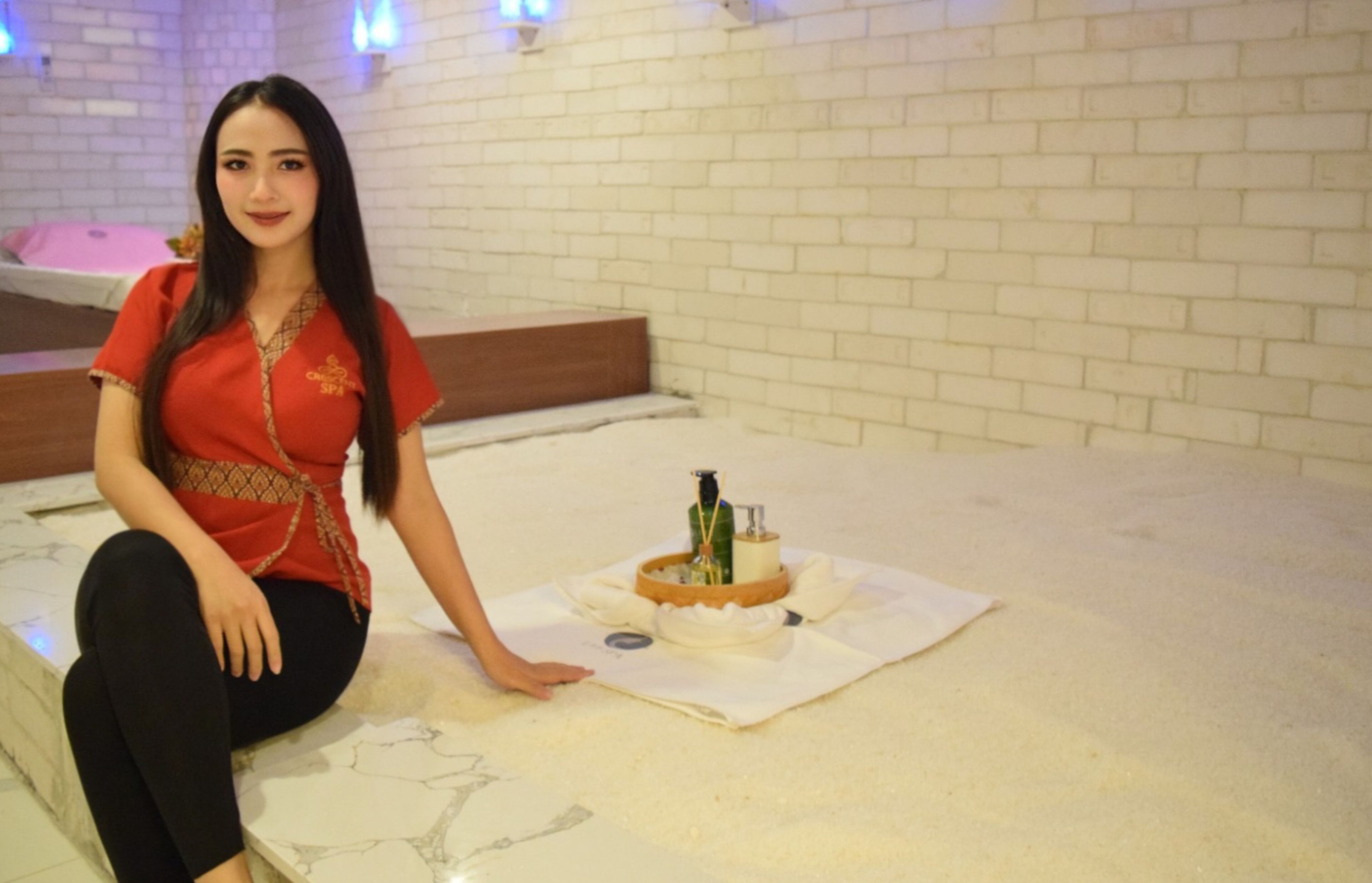 Une femme est assise sur un lit dans une pièce aux murs en briques blanches et dont la tête de lit est en bois.