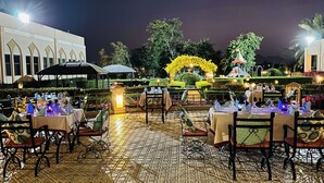 Un espace de restauration en plein air, avec des tables dressées pour les clients, entouré d’un jardin agrémenté de haies bien entretenues et d’une fontaine.