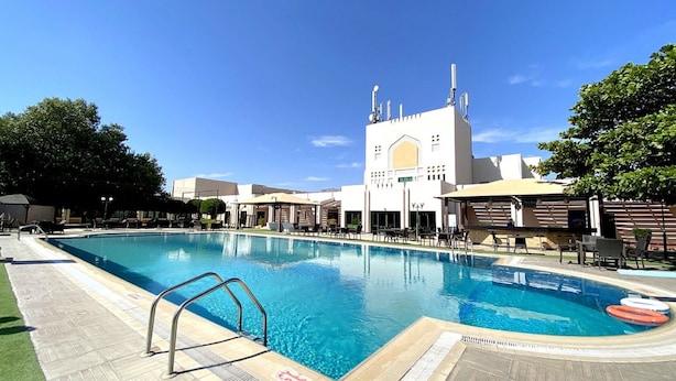 Une piscine dotée d’un poste de surveillance pour les sauveteurs, entourée de chaises longues et de parasols, avec un bâtiment en arrière-plan qui se distingue par une grande tour et des minarets.